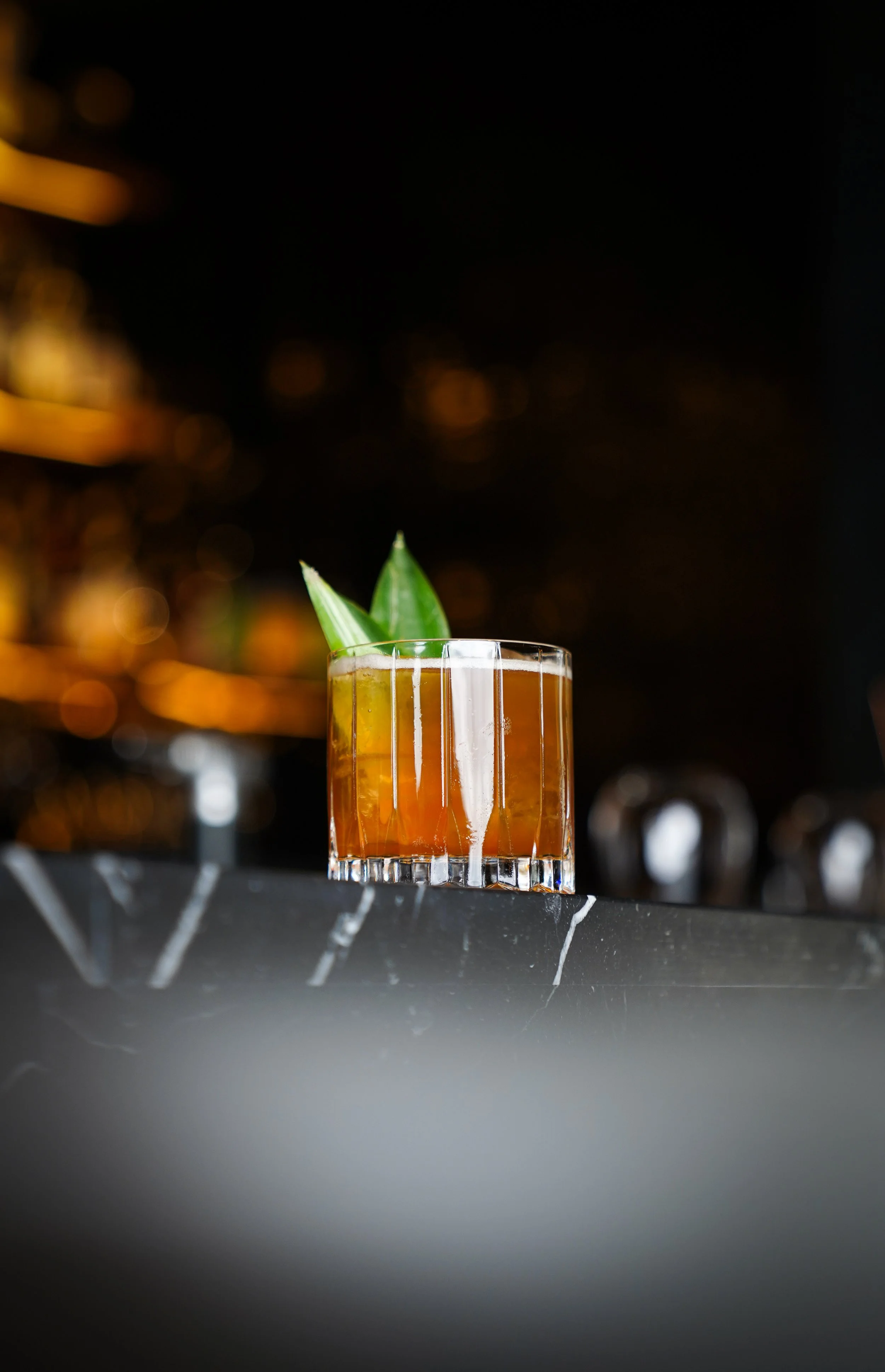 A cocktail in a glass garnished with pineapple leaves, placed on a dark bar counter with a blurred background.