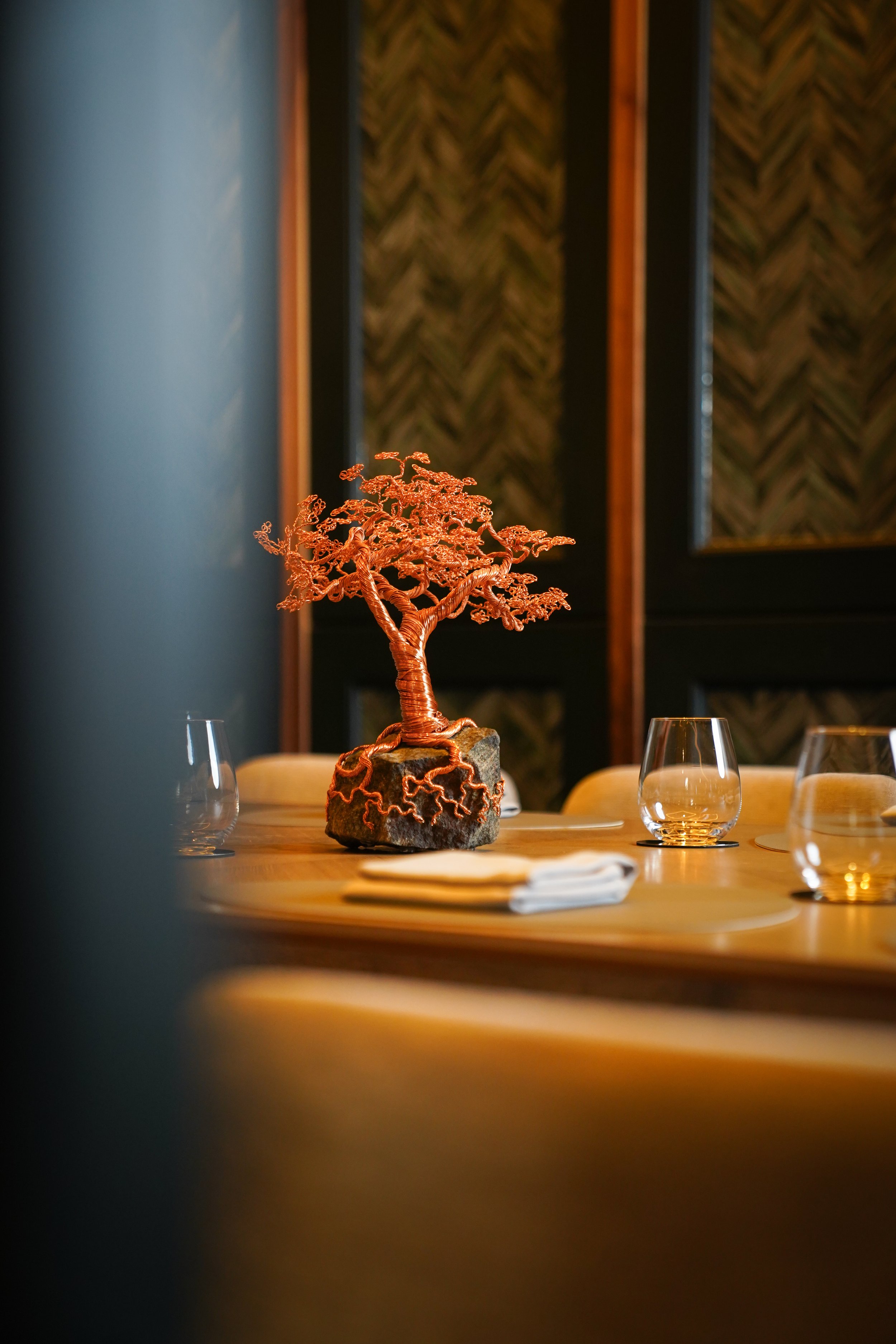 Copper wire sculpture of a bonsai tree on a dining table, surrounded by empty wine glasses and neatly folded napkins in a restaurant setting.
