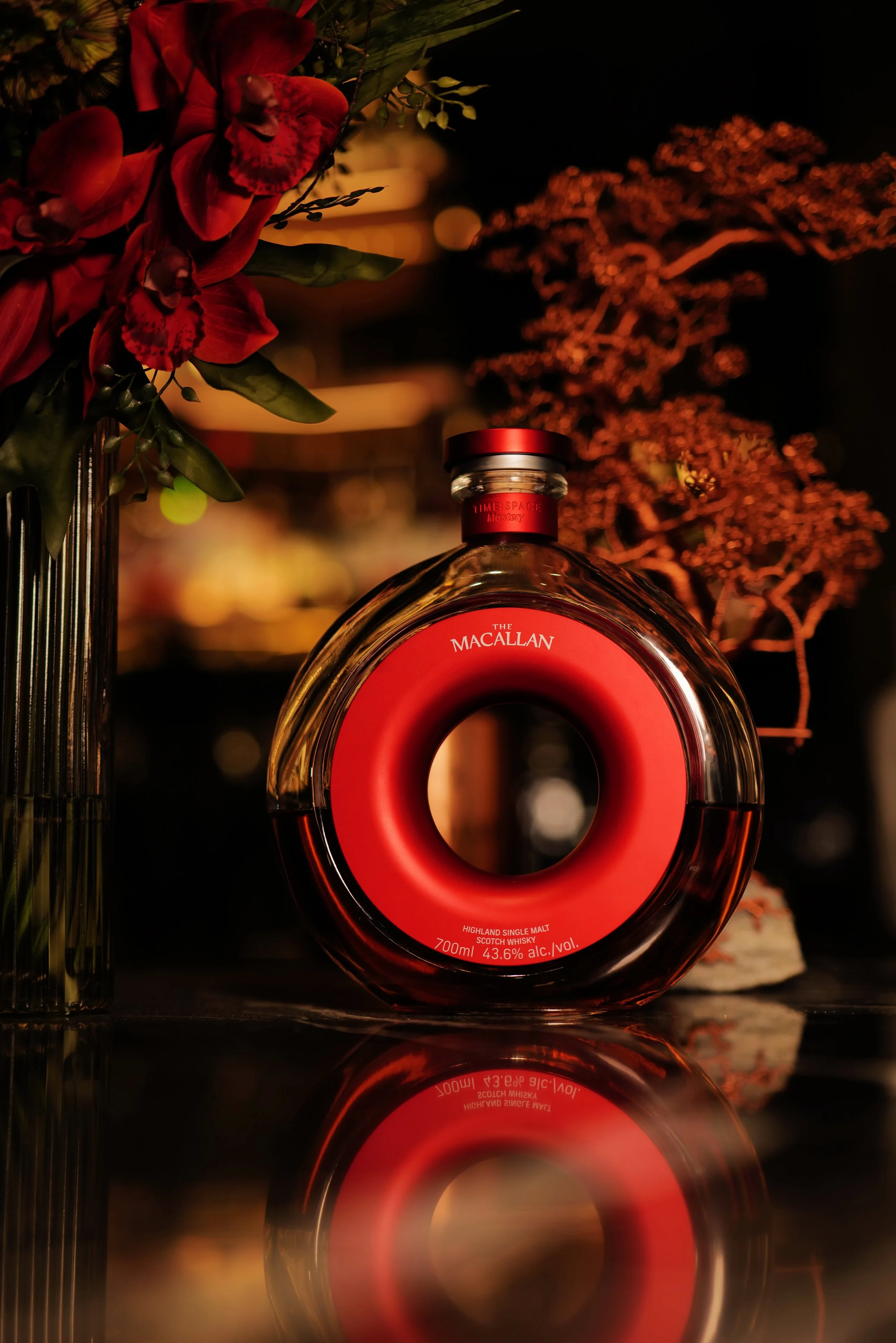 A round bottle of The Macallan Highland Single Malt Scotch Whisky with a red and black label and cap, set on a reflective surface with red and pink flowers and foliage in the background, creating a warm, elegant atmosphere.