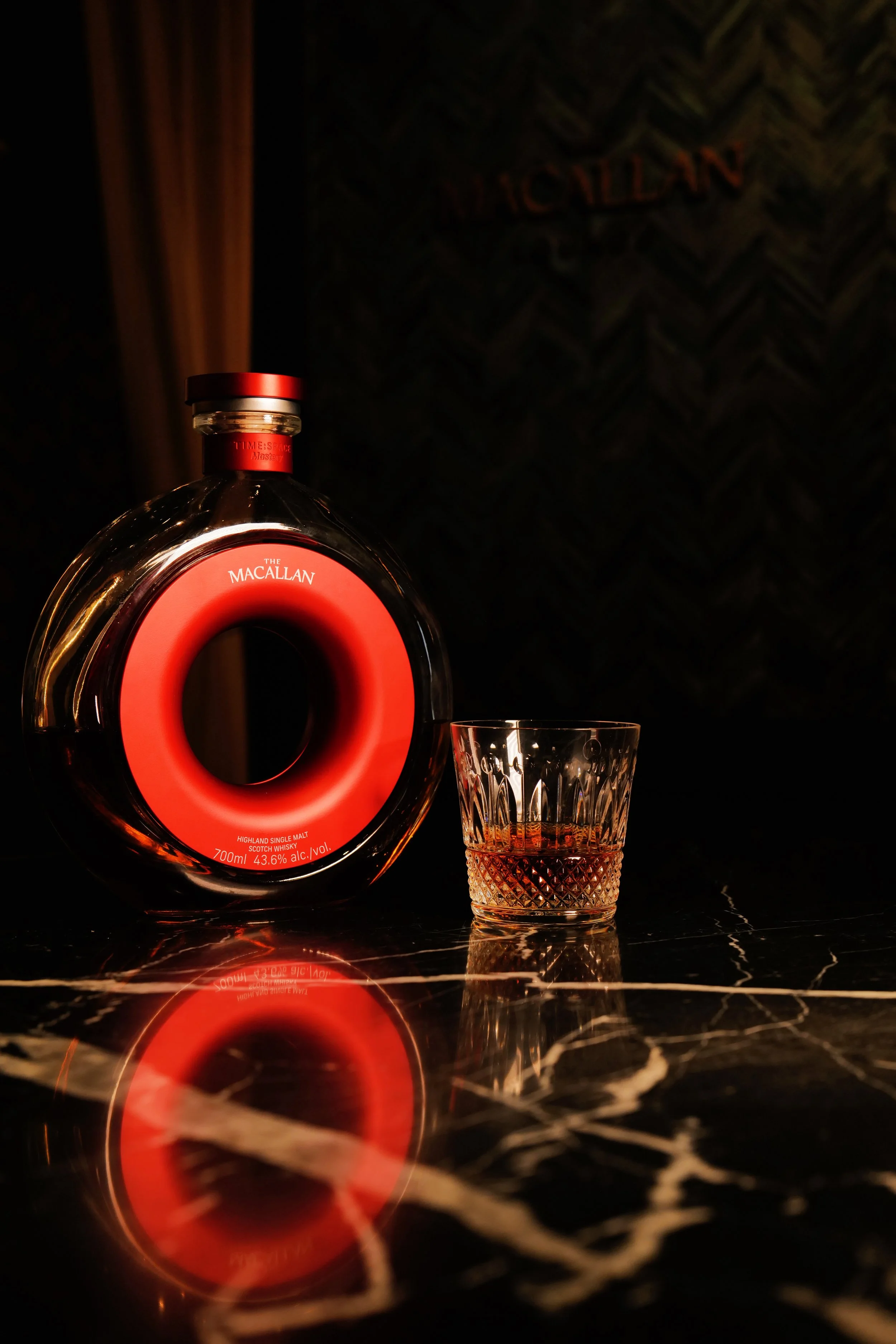 A bottle of The Macallan Highland Single Malt Scotch Whisky with a red circular design on the label, placed beside a glass of whisky on a reflective black marble surface.