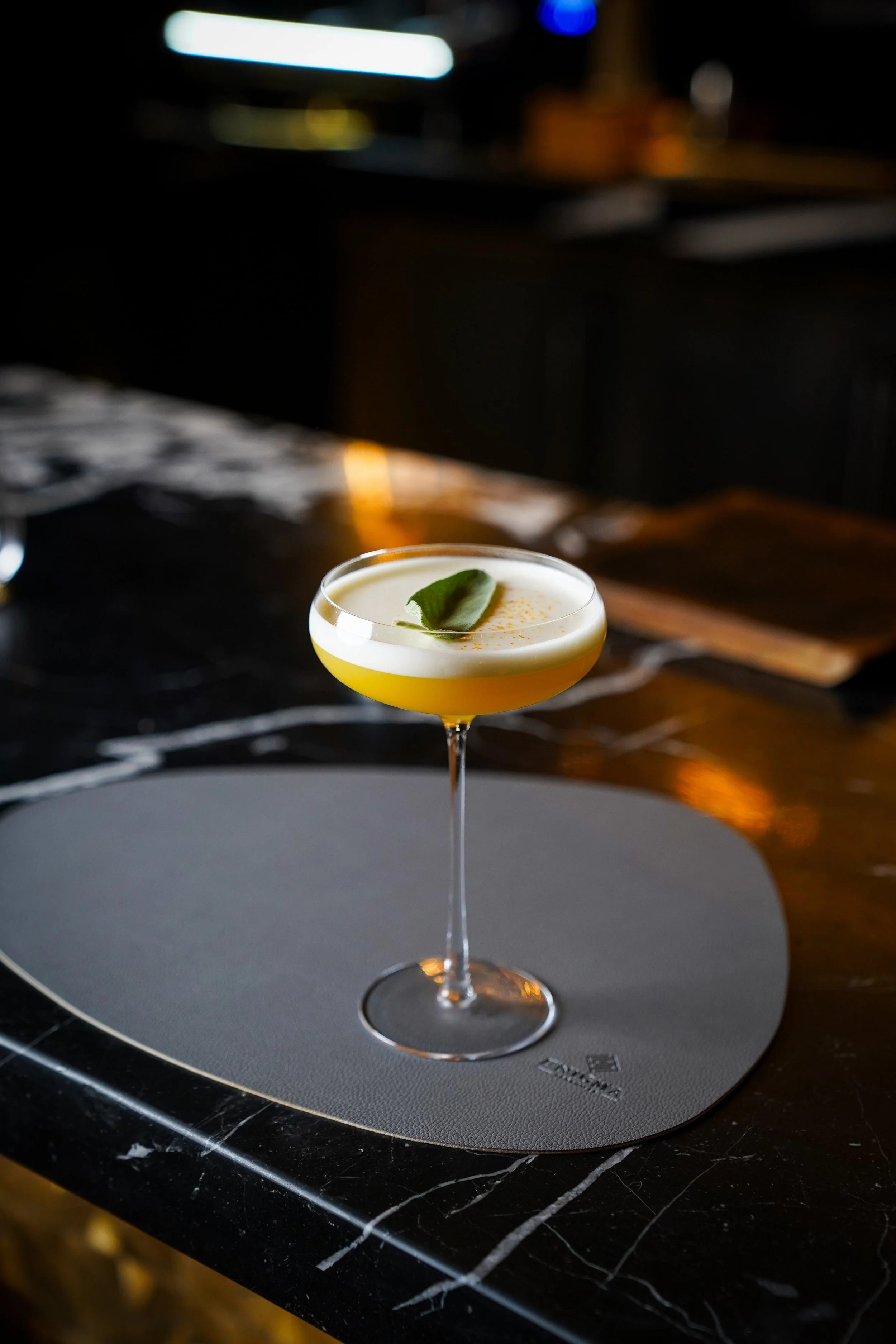 A cocktail in a stemmed glass with a white frothy top, garnished with a green leaf, placed on a gray mat on a dark marble countertop.
