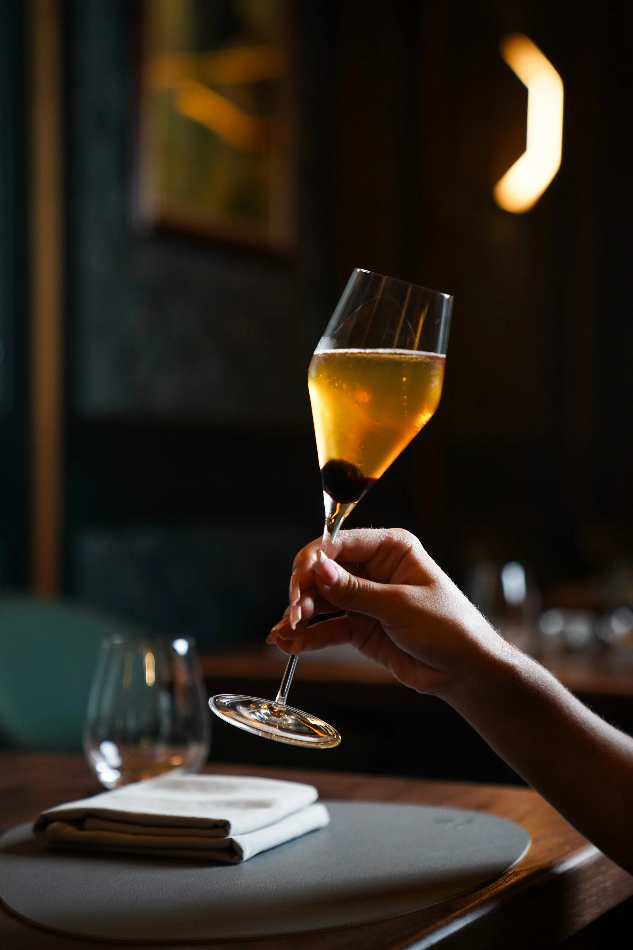 A hand holding a glass of champagne with a cherry at the bottom, set on a table in a dimly lit restaurant.