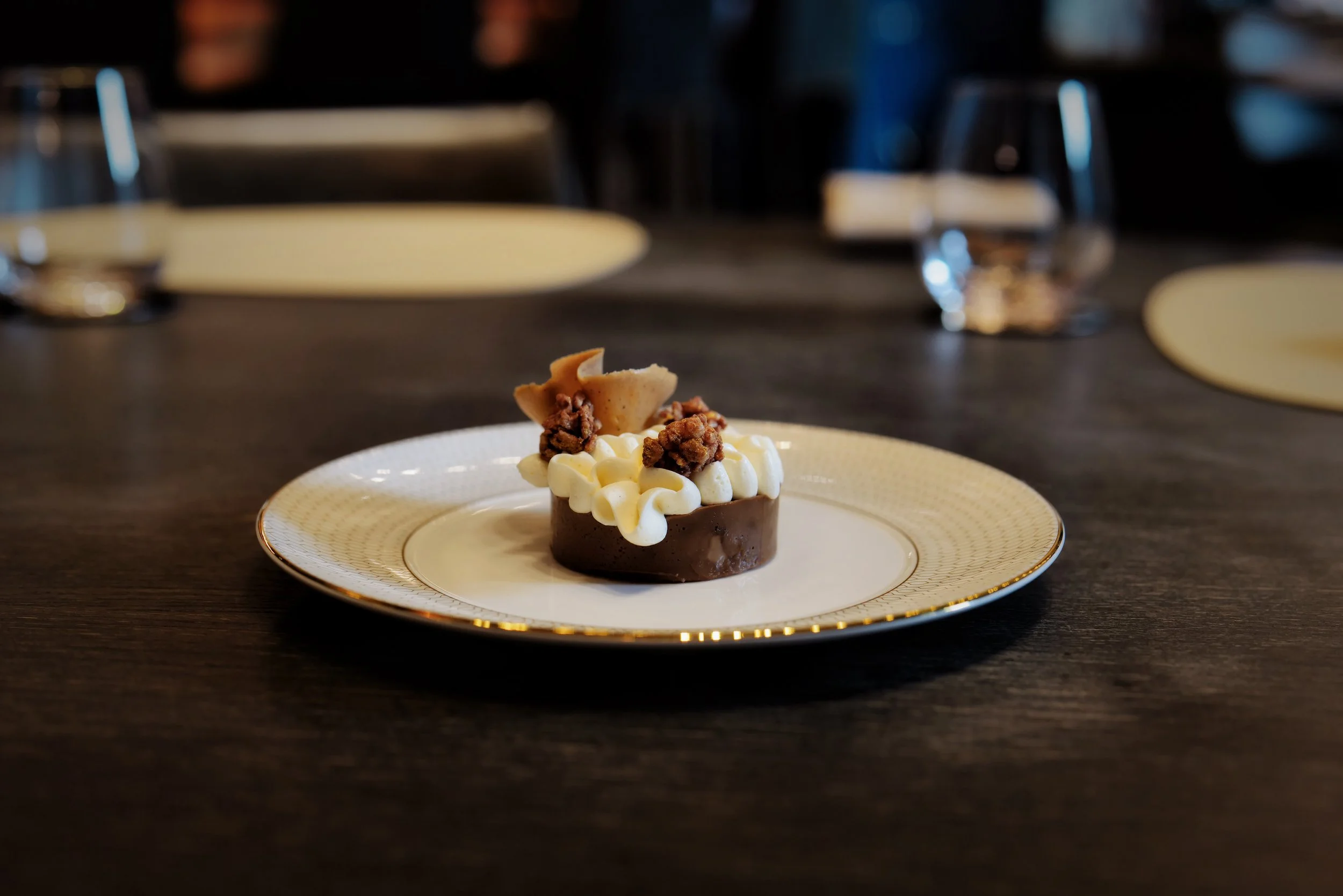 Elegant plated dessert featuring a chocolate mousse topped with decorative cream, crunchy toppings, and cookie garnish, on a white china plate with gold trim, set on a dark wooden table in a dimly lit restaurant.