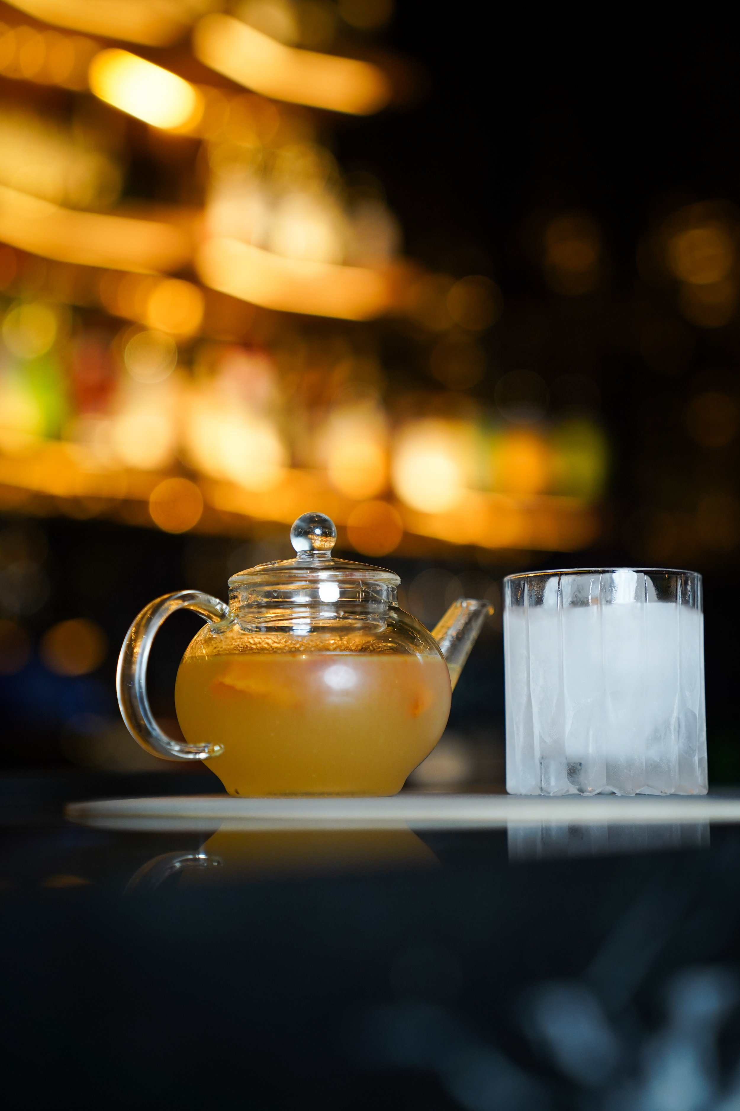 A glass teapot filled with a light-colored liquid, next to a glass with ice cubes on a white plate, with a warm, out-of-focus background of shelves with bottles and glasses.