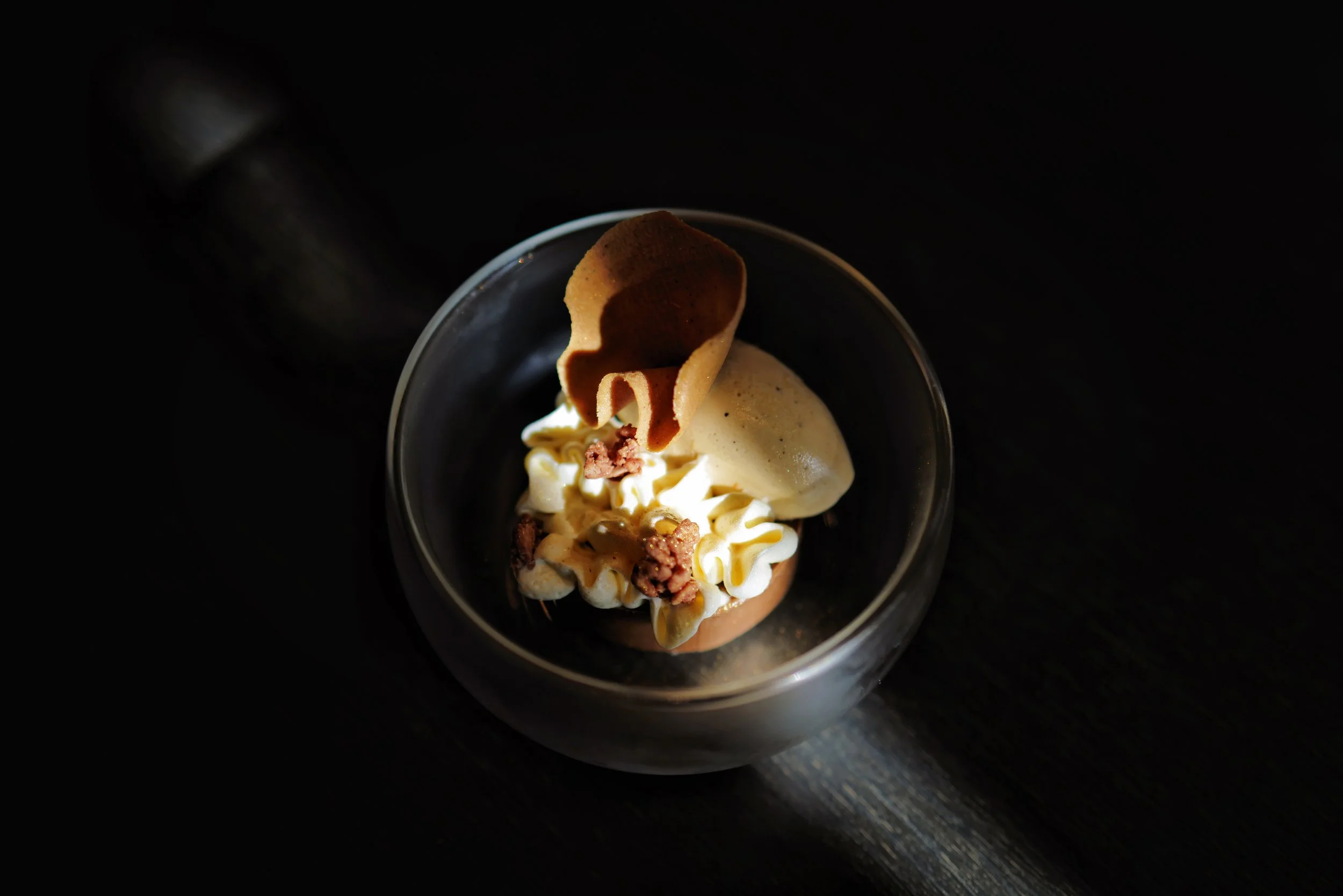 A dessert in a glass bowl featuring a scoop of vanilla ice cream, whipped cream, a decorative tuile cookie, and small crumbles on top, set against a dark background.