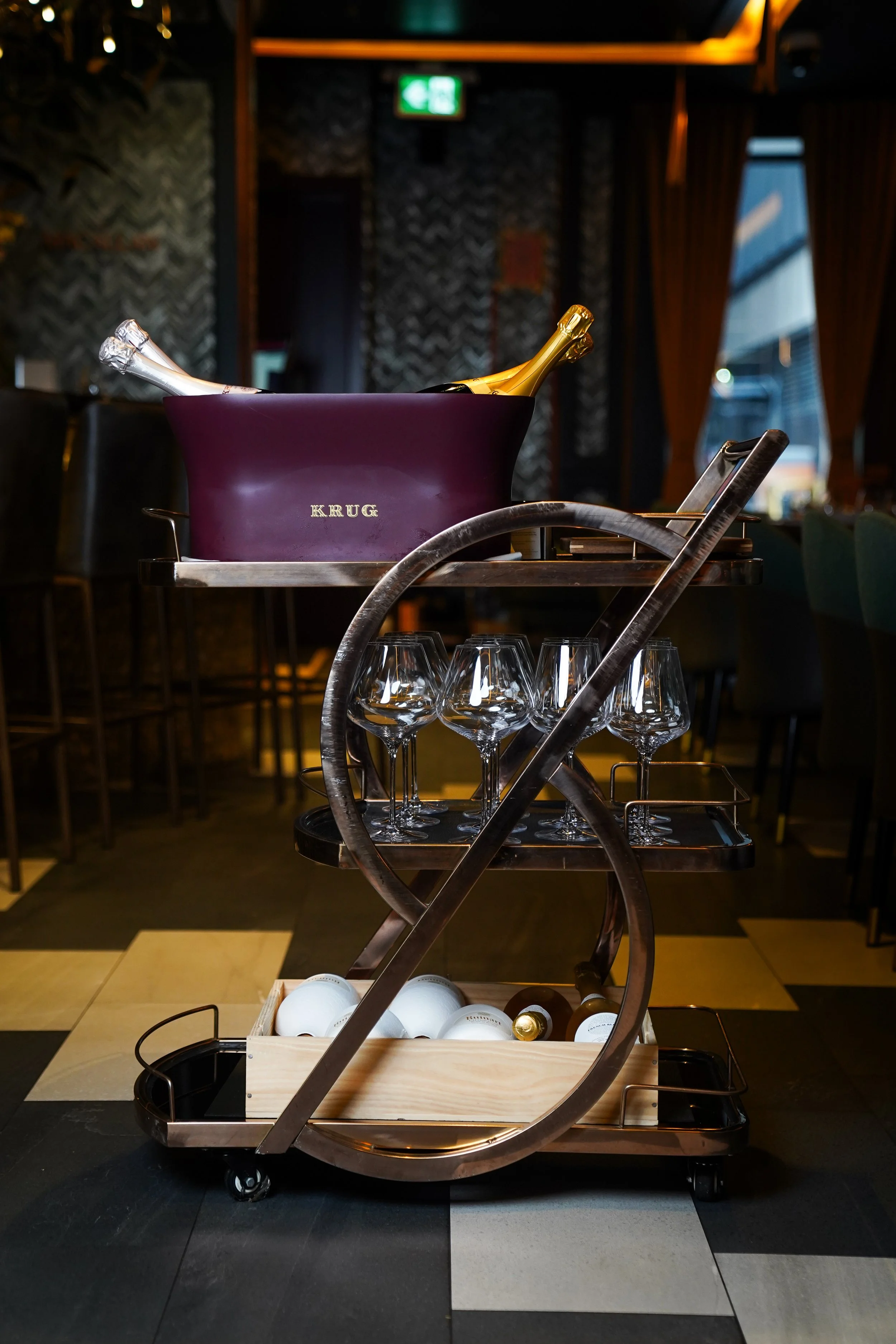A hotel room service cart with champagne bottles in an ice bucket, empty wine glasses, and a small wooden box with white and brown bottles inside, in a dimly lit restaurant or bar.