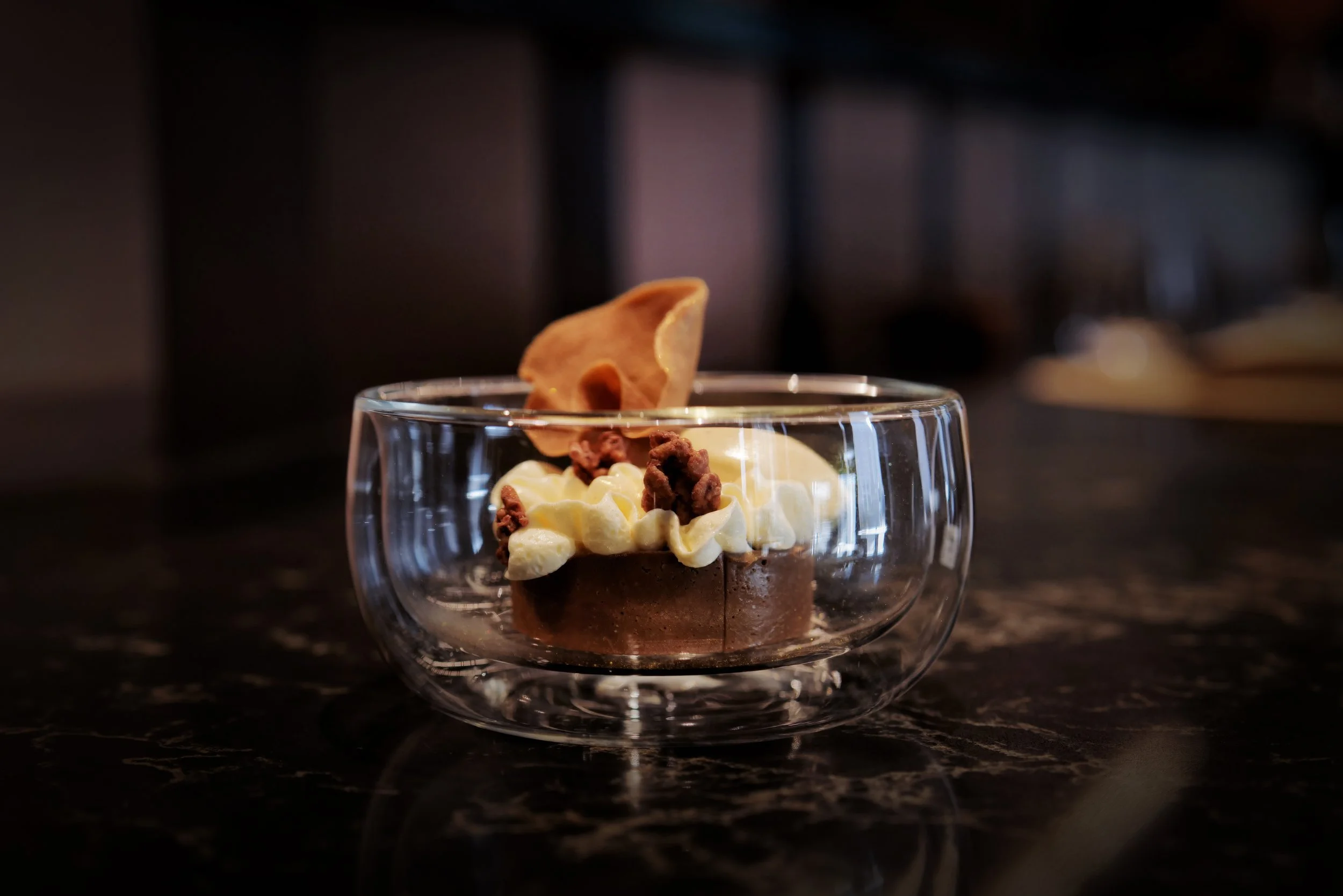 A small chocolate dessert topped with whipped cream, chocolate crumbles, and a cookie, served in a clear glass bowl on a dark marble surface.