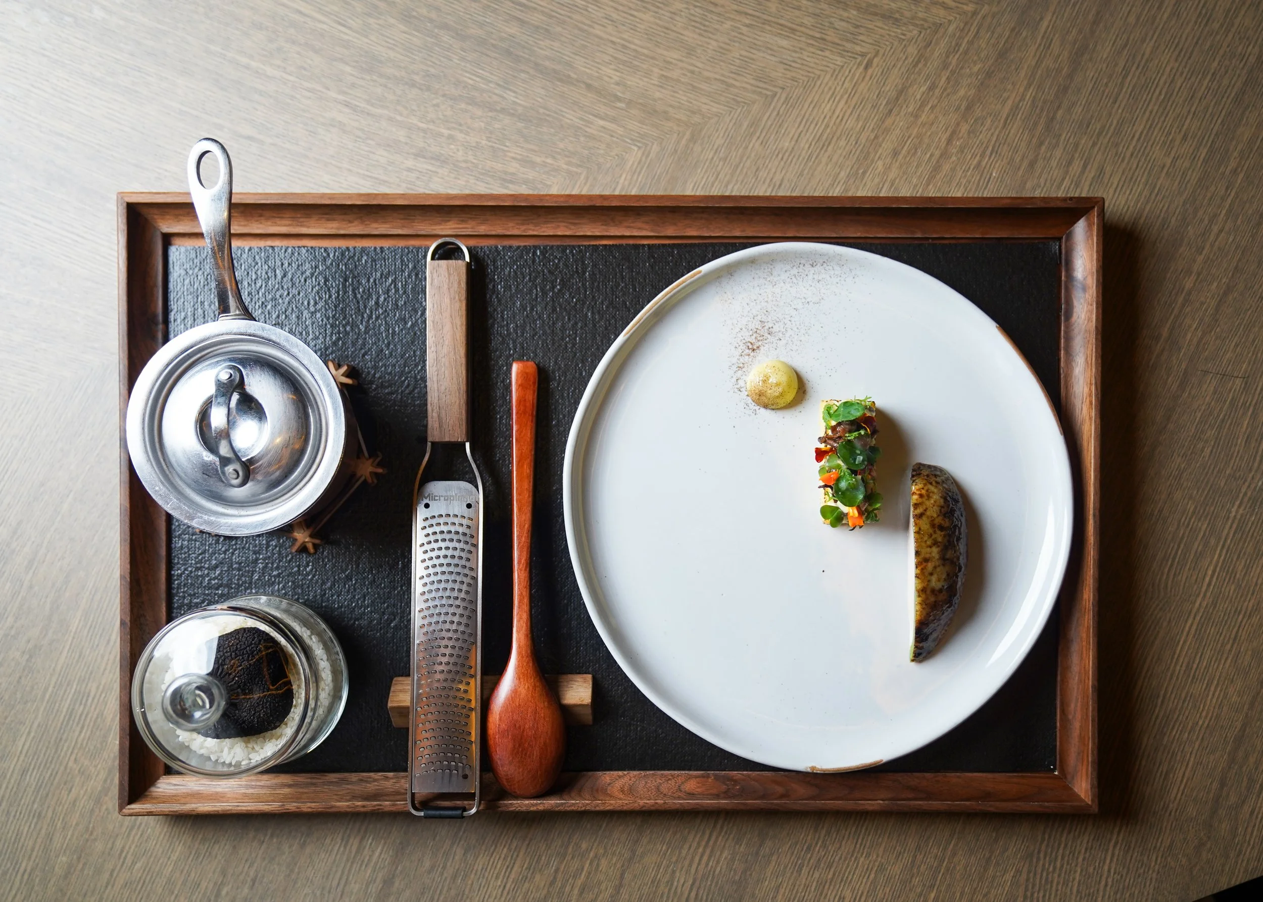 Empty white plate with small food portions, a yellow ball, and a colorful garnish on a wooden tray with utensils and containers around, seen from above.
