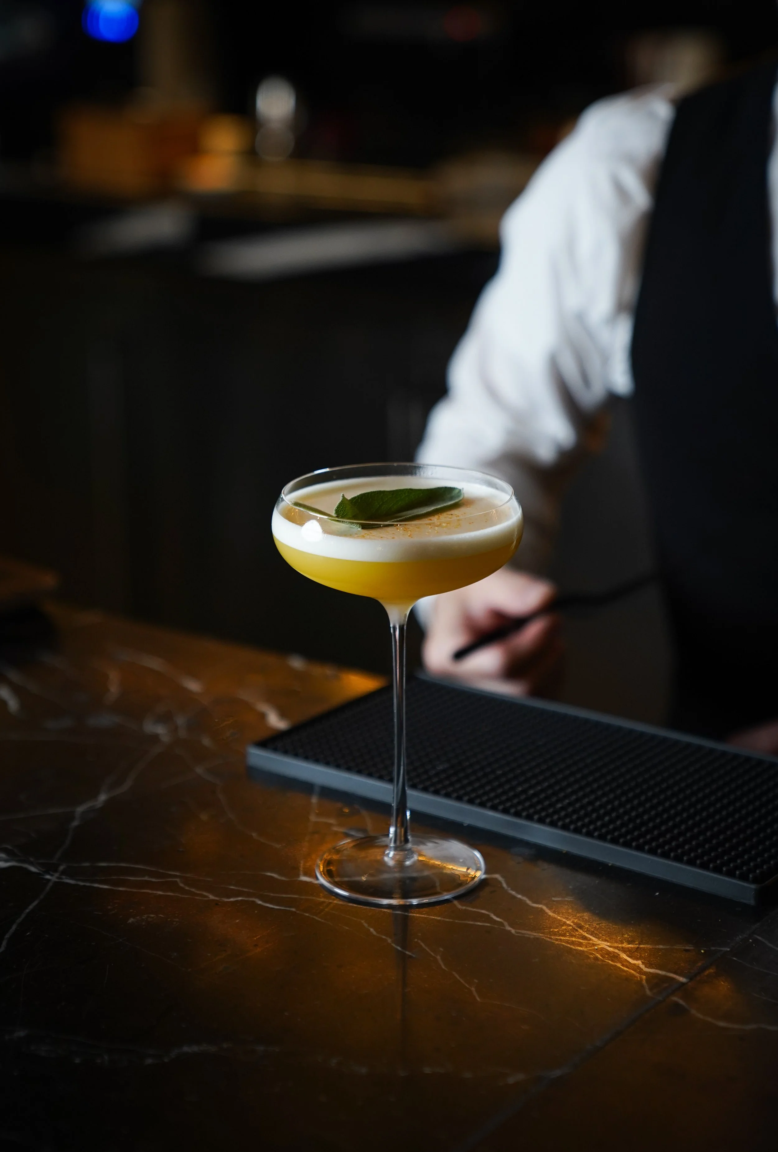 A yellow cocktail with a lime garnish on a cocktail glass on a dark marble bar counter.