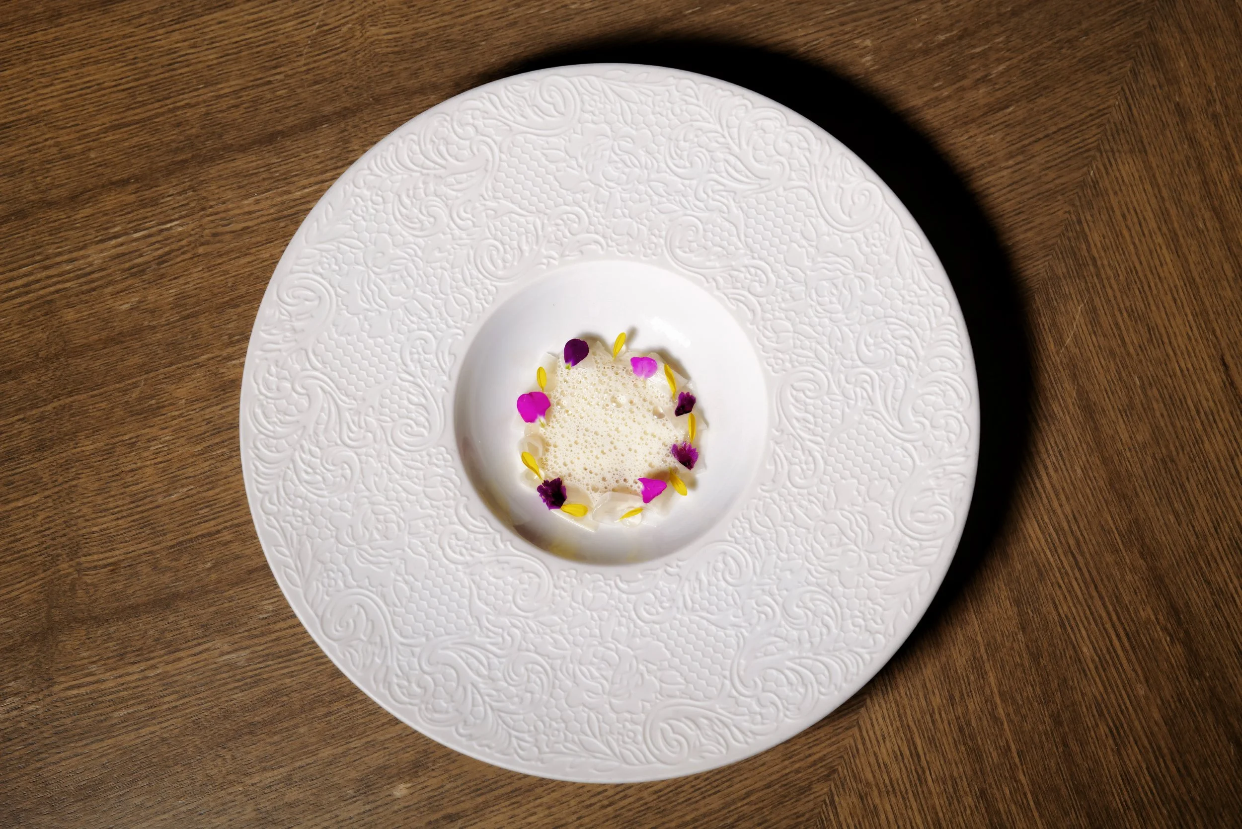 Elegant white textured plate with a small foam dish garnished with purple and yellow edible flowers, placed on a wooden surface.