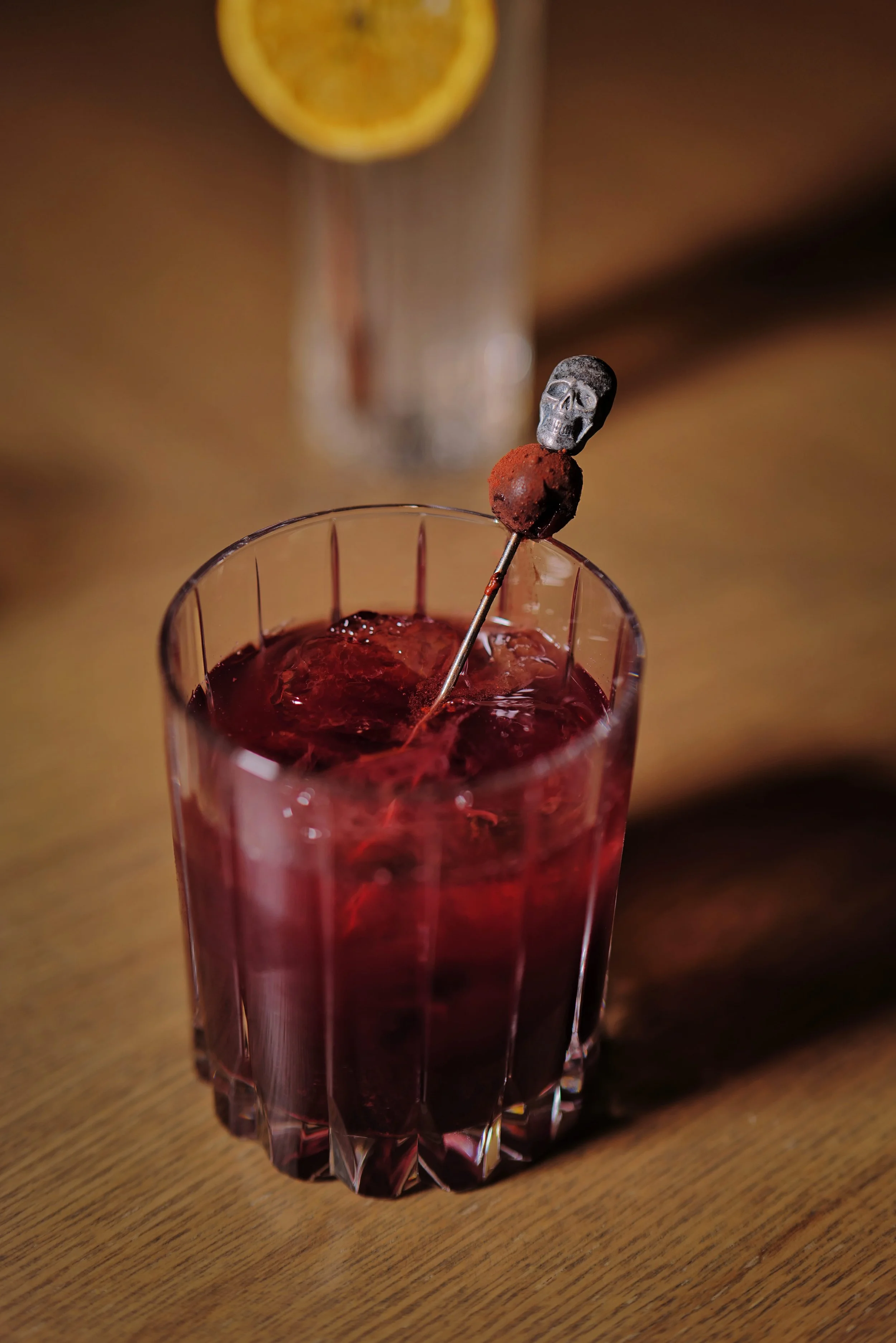 A close-up of a glass of dark red cocktail with a skull-shaped pick inserted into a piece of fruit, on a wooden surface with a lemon slice in the background.