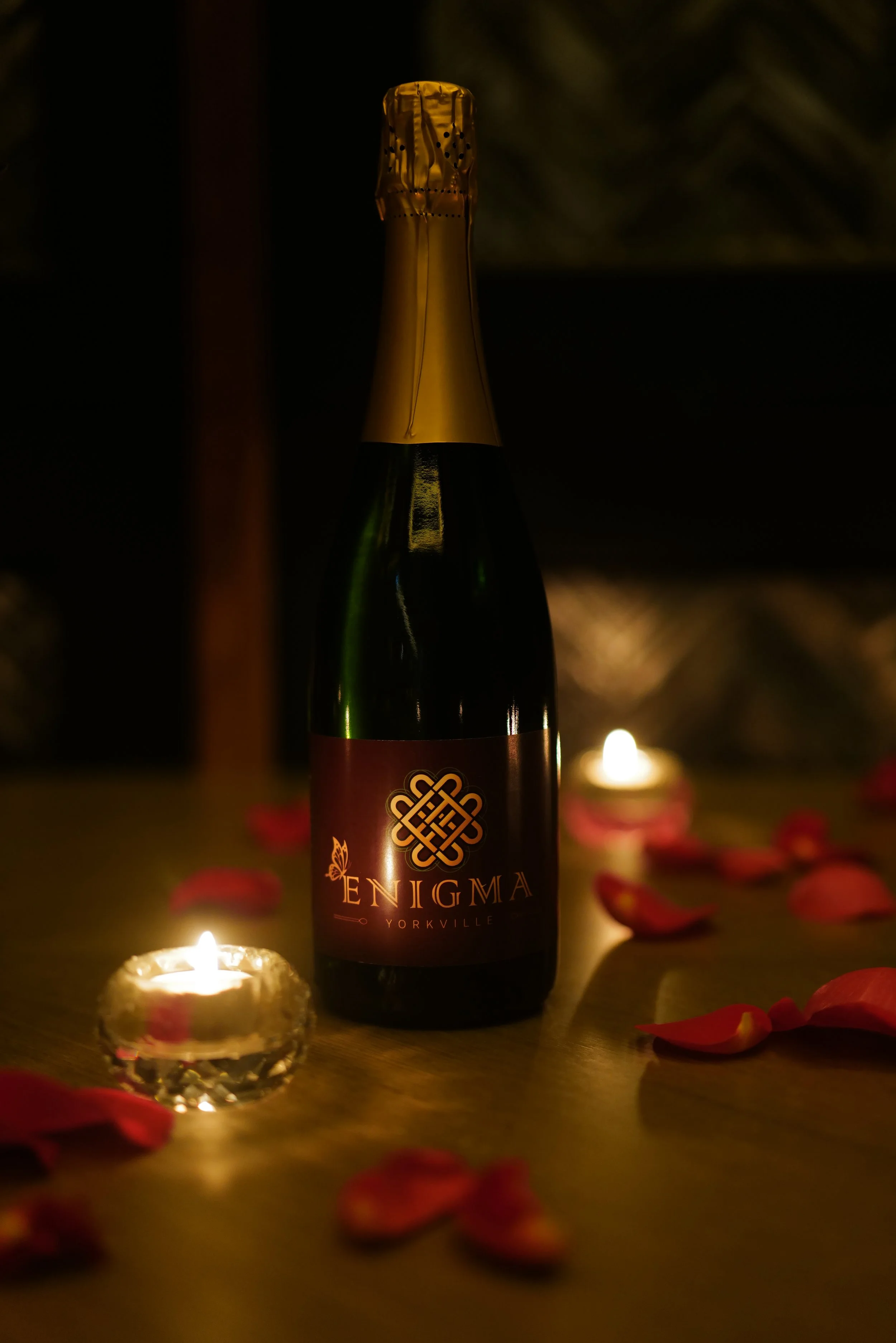 A bottle of Enigma Yorkville sparkling wine on a table, surrounded by scattered pink rose petals and lit candles in small glass holders, in a dimly lit setting.