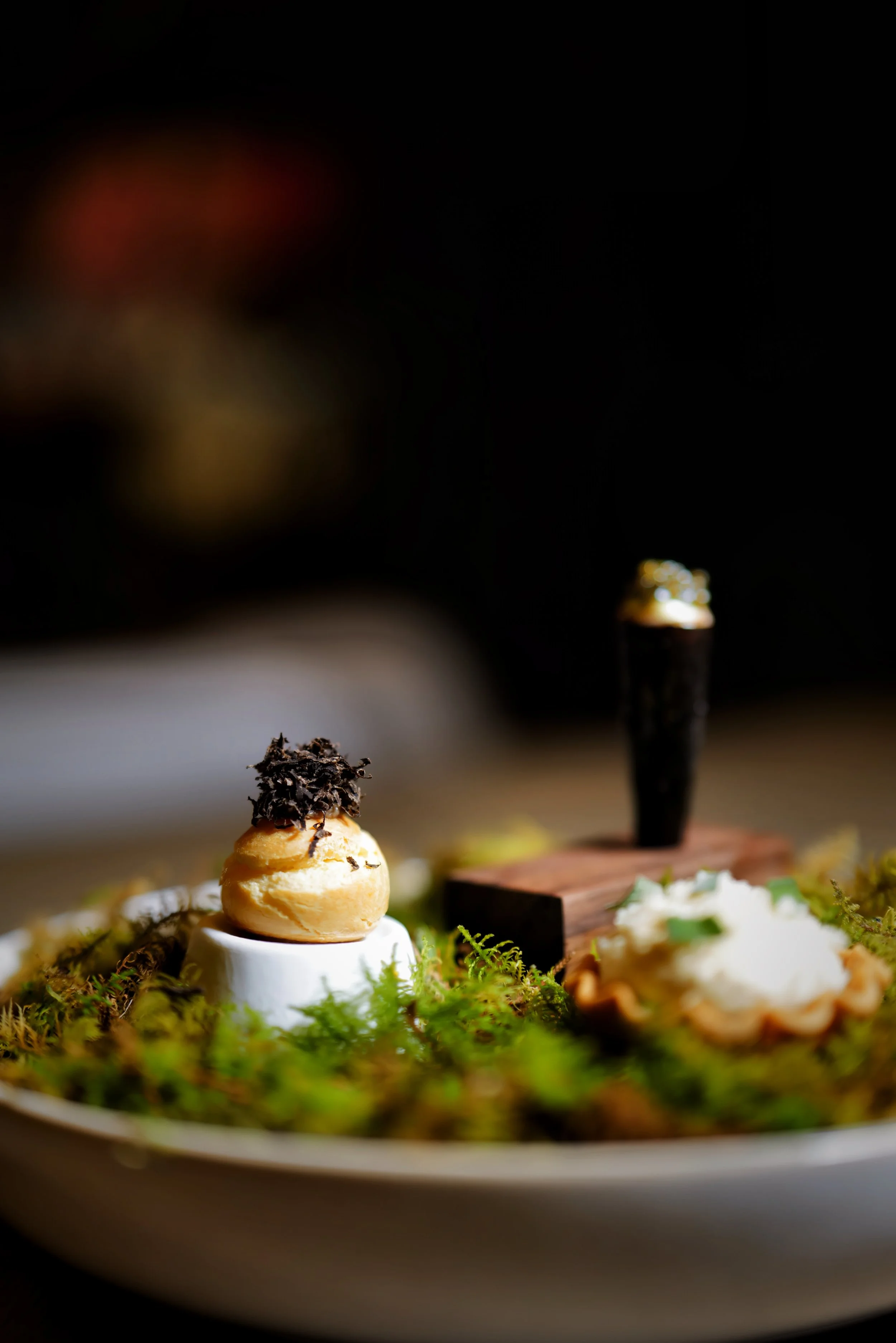 A dish with a small pastry topped with dark shredded garnish on a white cup, placed on a bed of greenery, with other appetizers on the plate, out of focus.