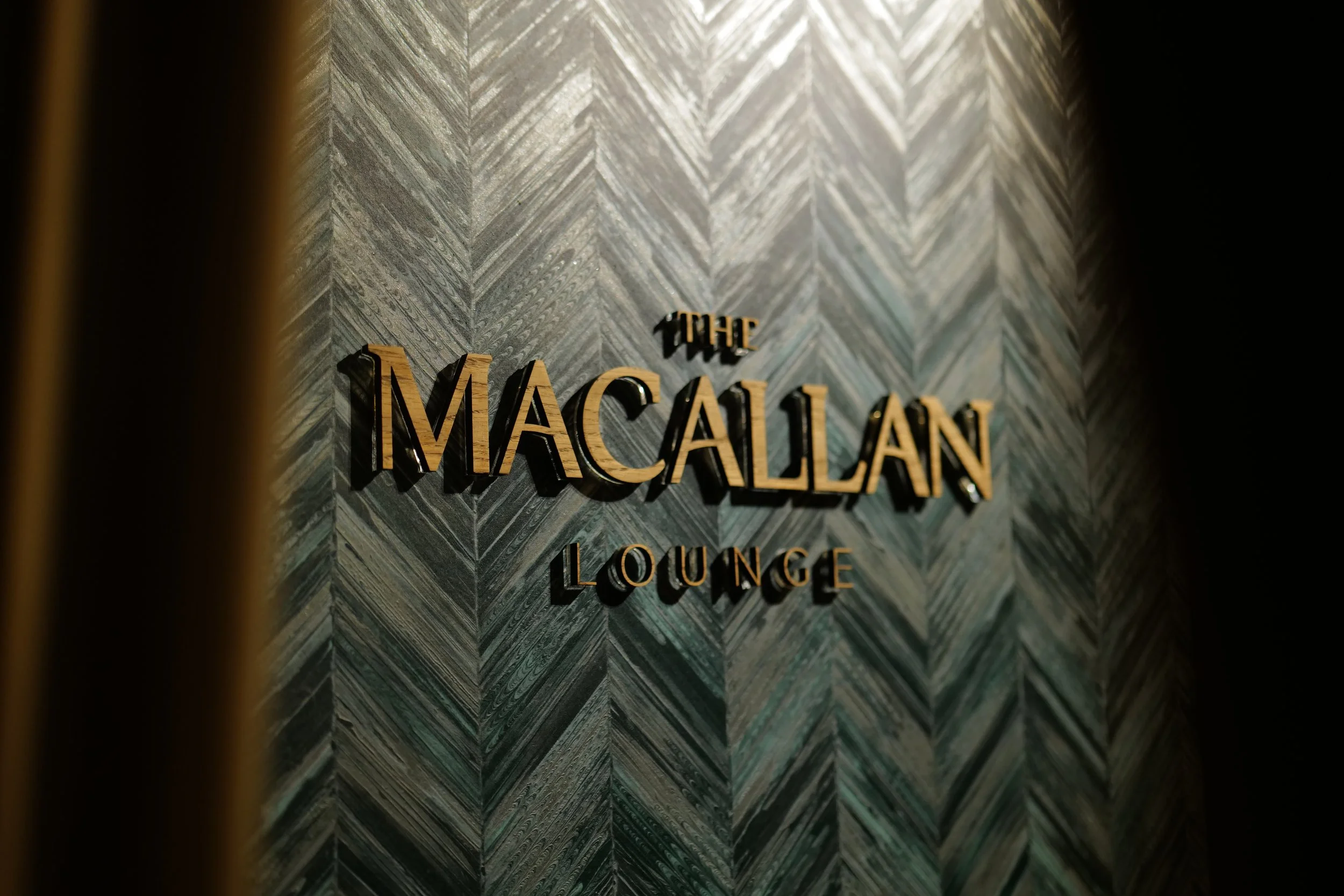 Sign on a textured gray wall reading 'The Macallan Lounge' in gold lettering.