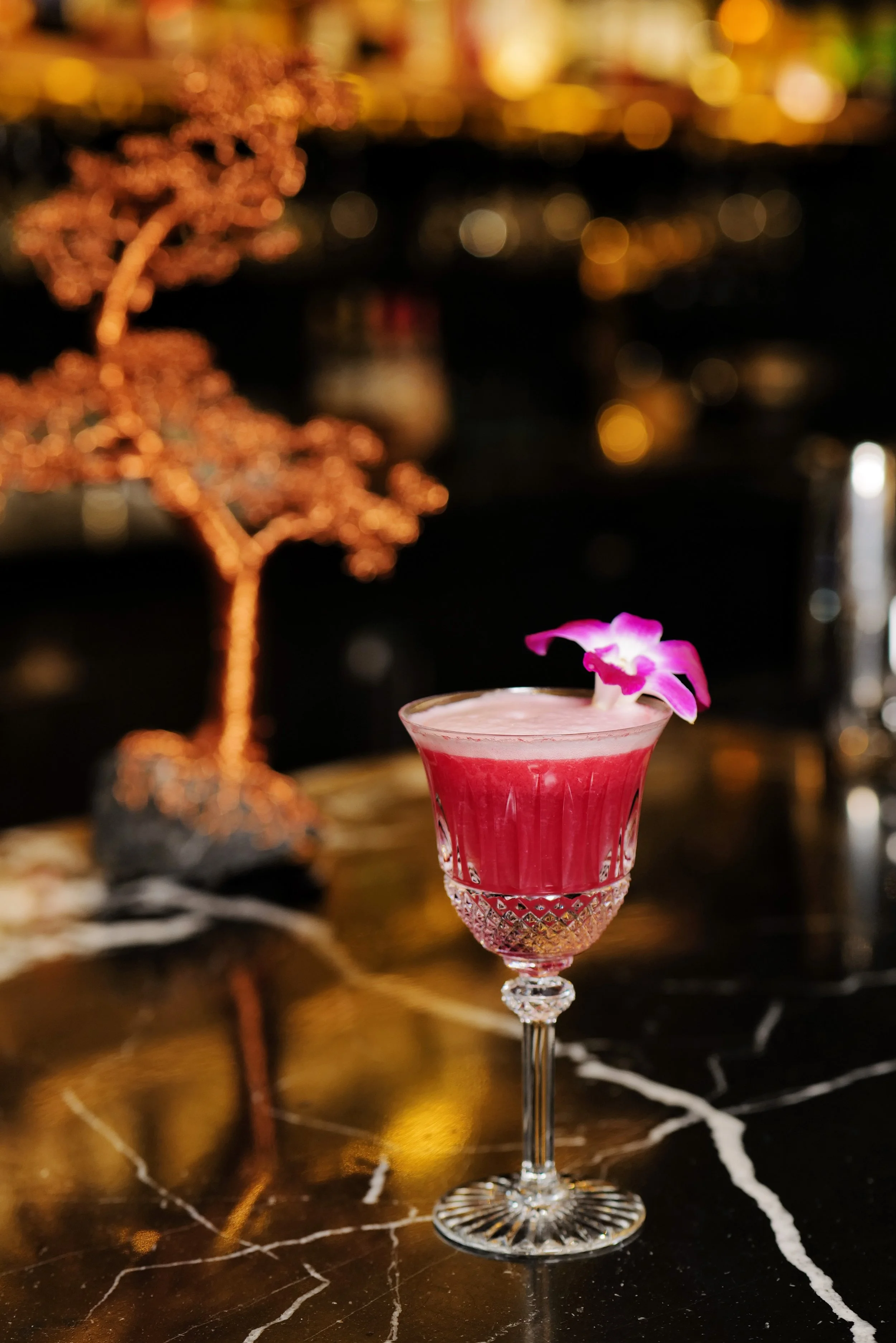A pink cocktail garnished with a pink flower in a crystal glass on a dark marble bar counter.