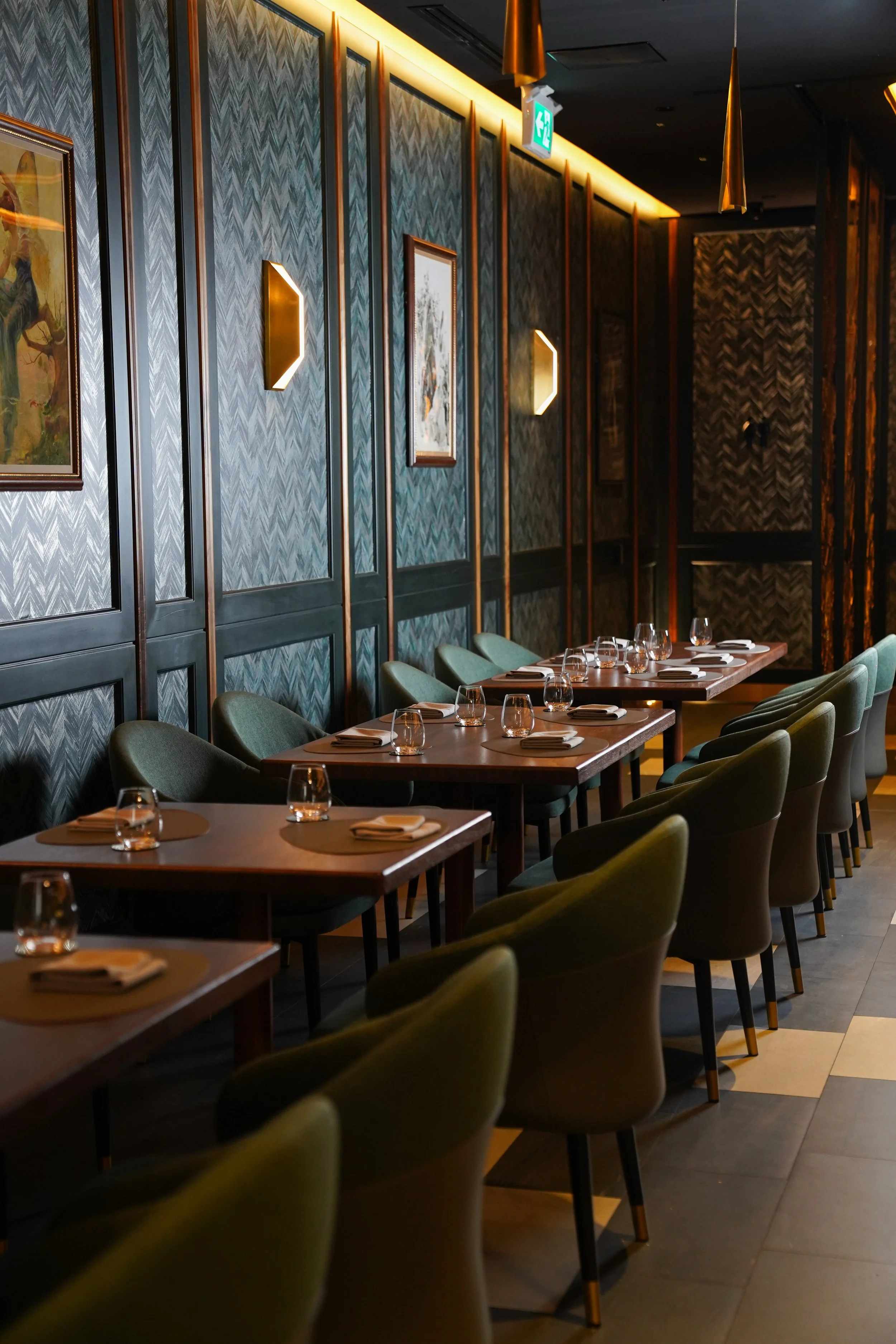 Empty restaurant with tables set with glassware and napkins, green chairs, wall with framed paintings and wall lights, and an illuminated exit sign.