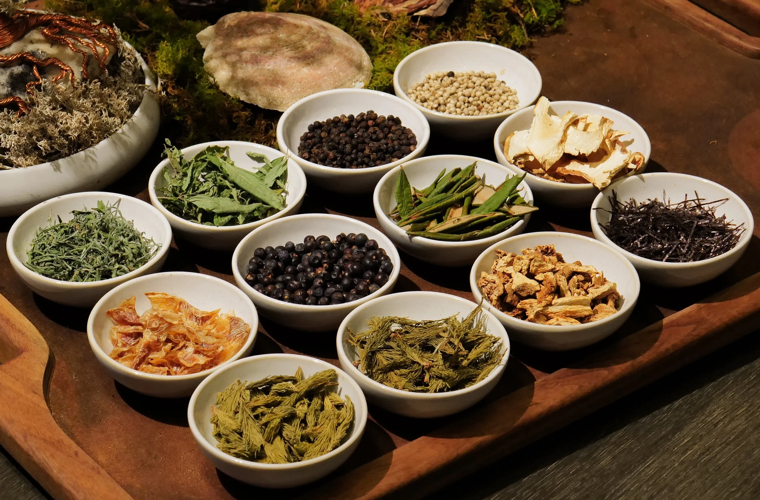 A wooden tray with various bowls containing herbs, spices, and dried plants, including green leaves, black and white peppercorns, dried roots, and other botanical ingredients, with a decorative rock and moss in the background.