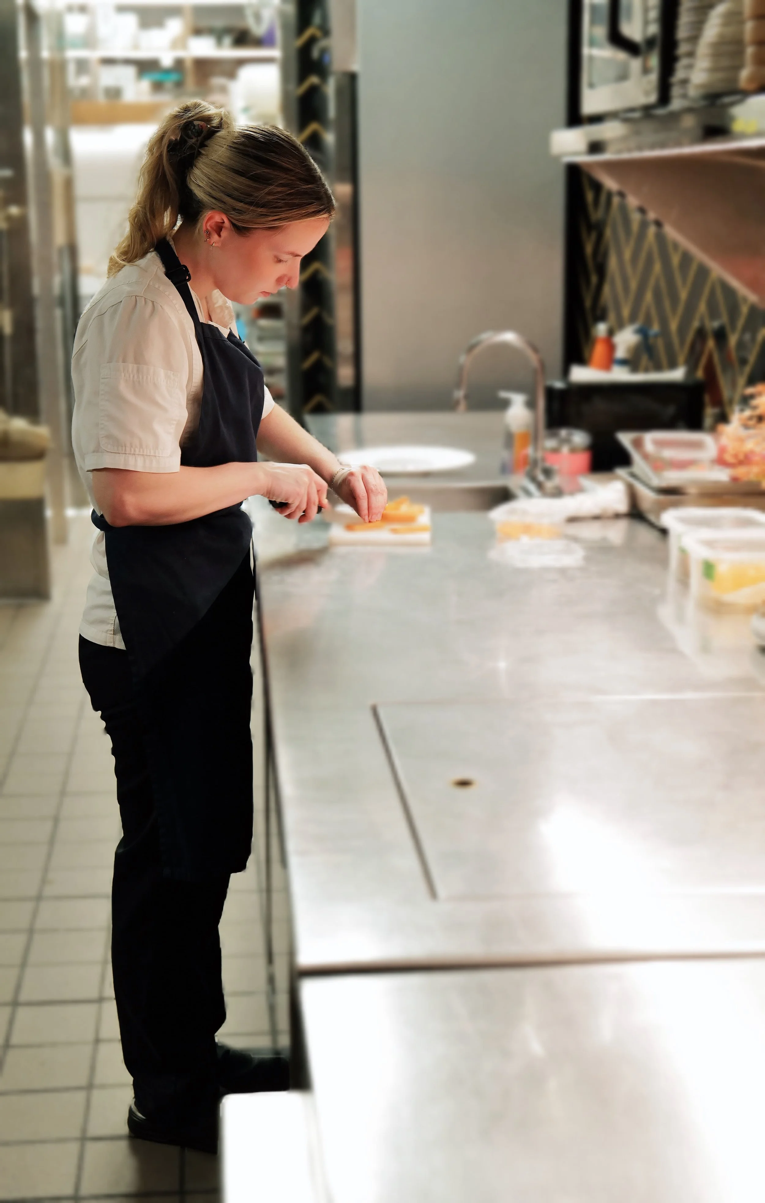 A woman in a beige shirt and black apron preparing food in a commercial kitchen.