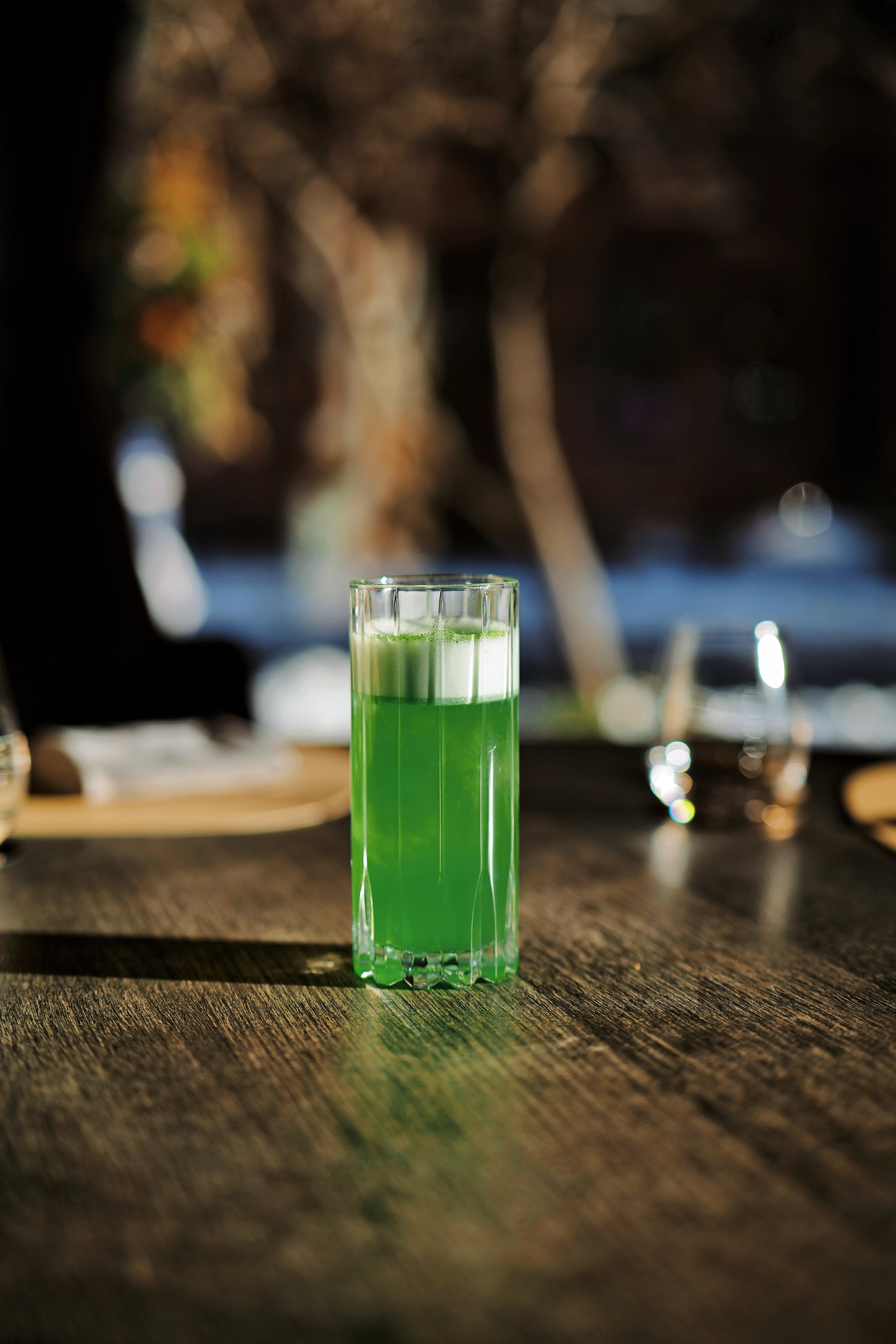 A tall glass filled with green liquid on a wooden table, with a blurred outdoor background.