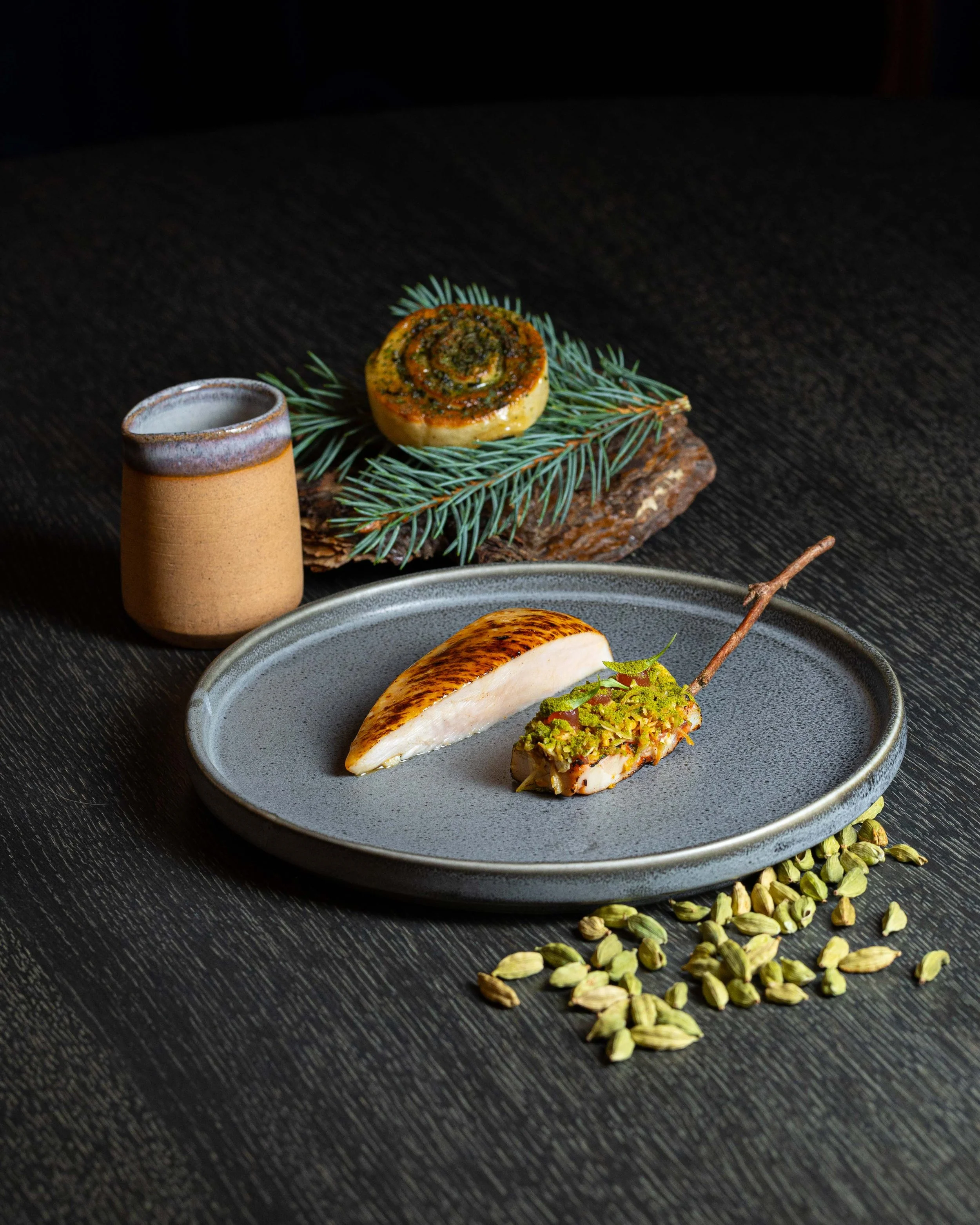 A plated gourmet meal with a seared piece of chicken breast and a small, herb-topped fillet, garnished with herbs and seeds, on a gray plate. In the background, a round roasted vegetable and a sprig of pine with additional seeds scattered on the blac