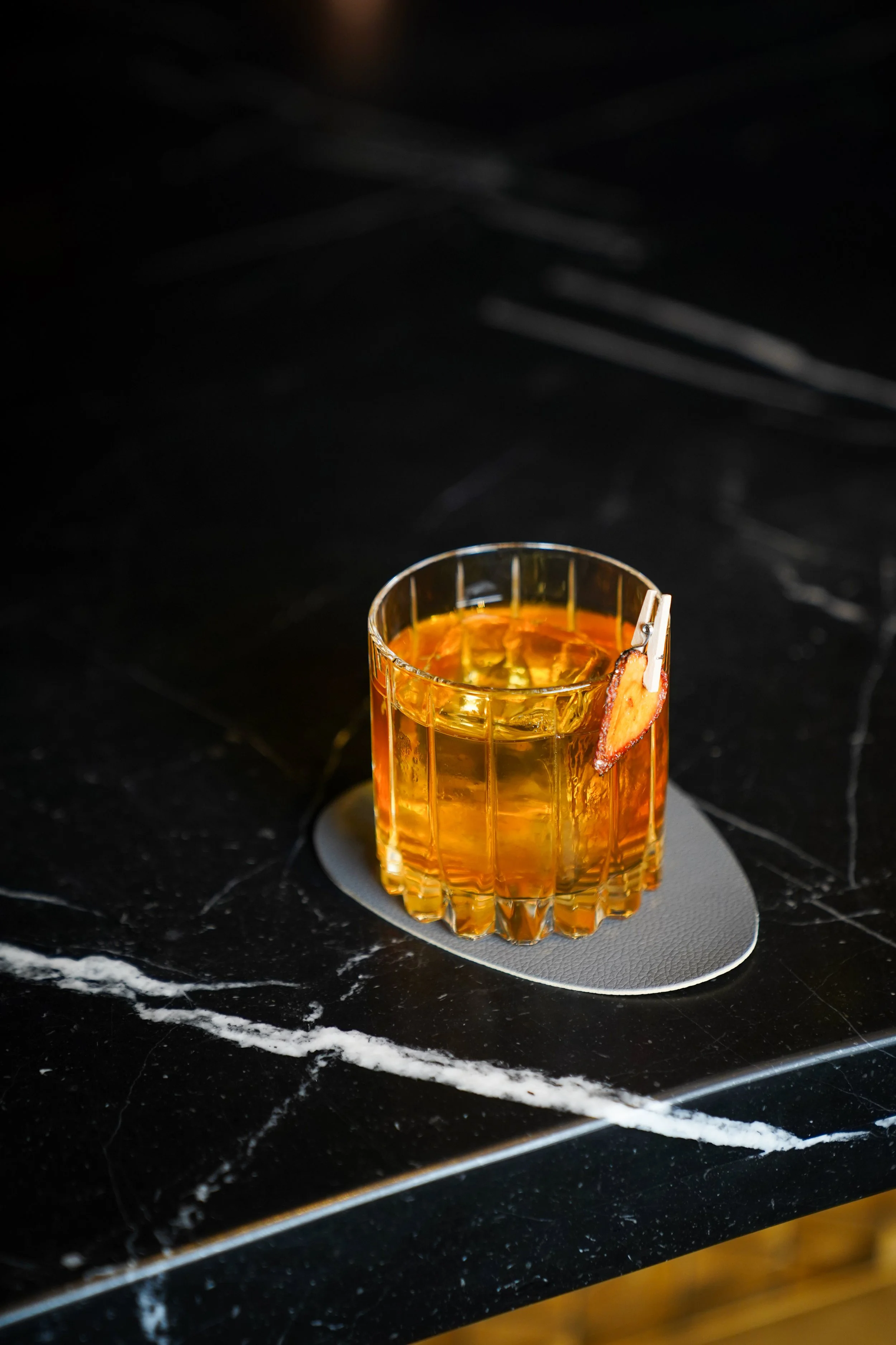 A glass of whiskey with ice on a black marble countertop, garnished with a small piece of bacon clipped on the rim.