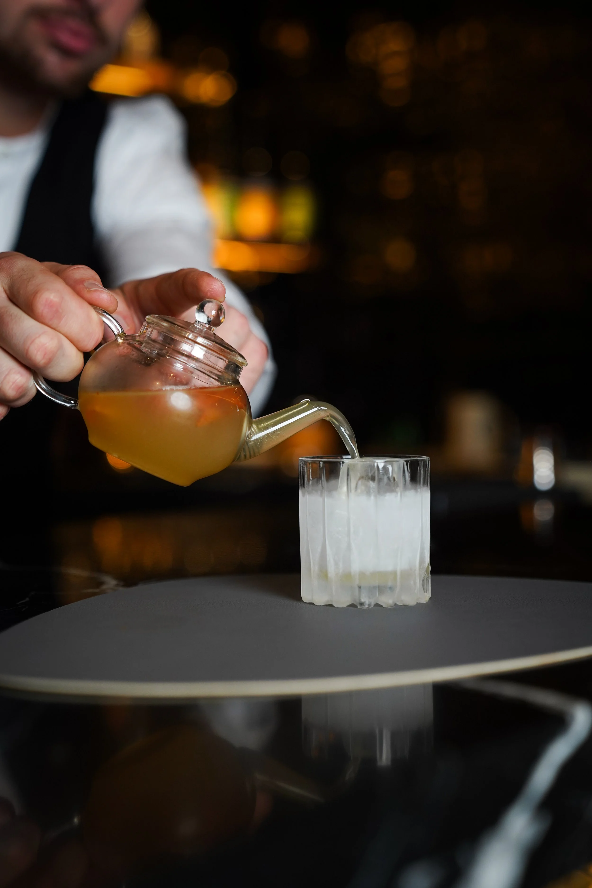 A bartender pours a drink from a teapot-shaped vessel into a glass at a bar with a blurred background.