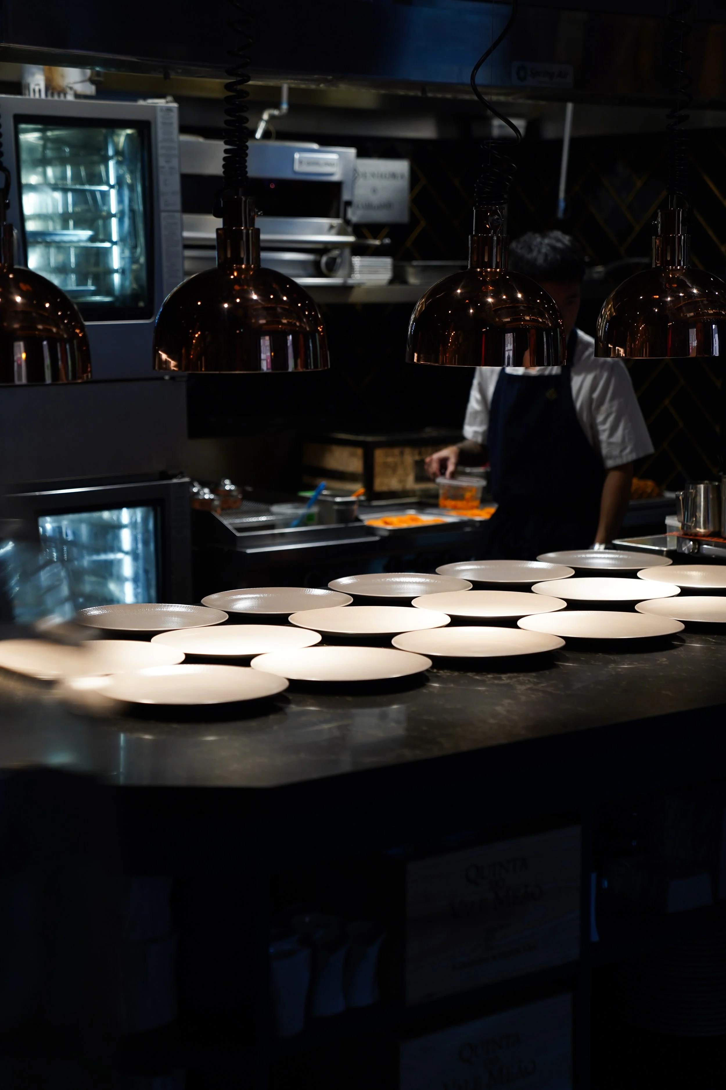 Empty white plates arranged on a dark restaurant counter with a chef working in the background under hanging copper pendant lights.