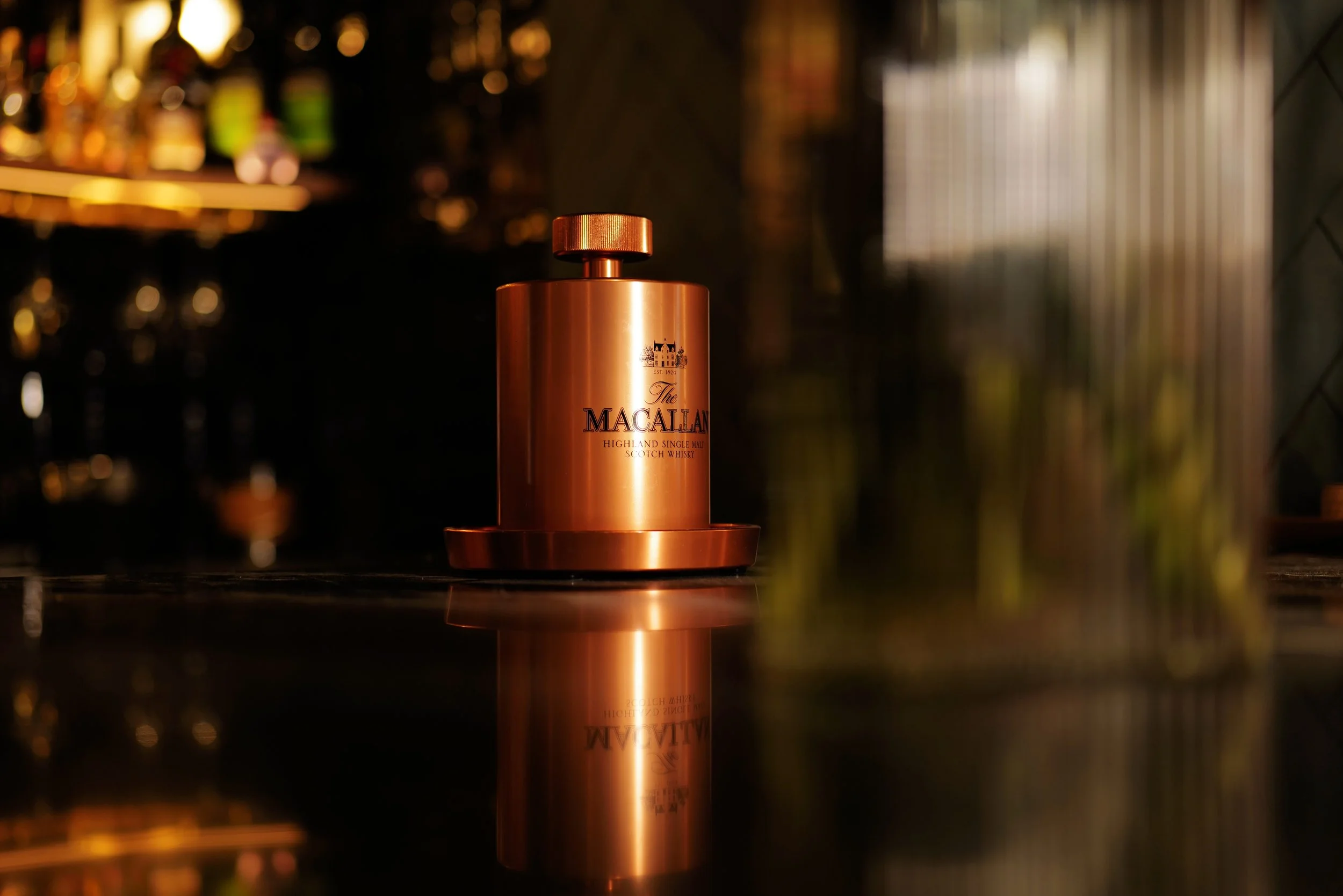 A copper-colored bottle of The Macallan Highland Single Malt Scotch Whisky on a reflective surface with a blurred bar background.