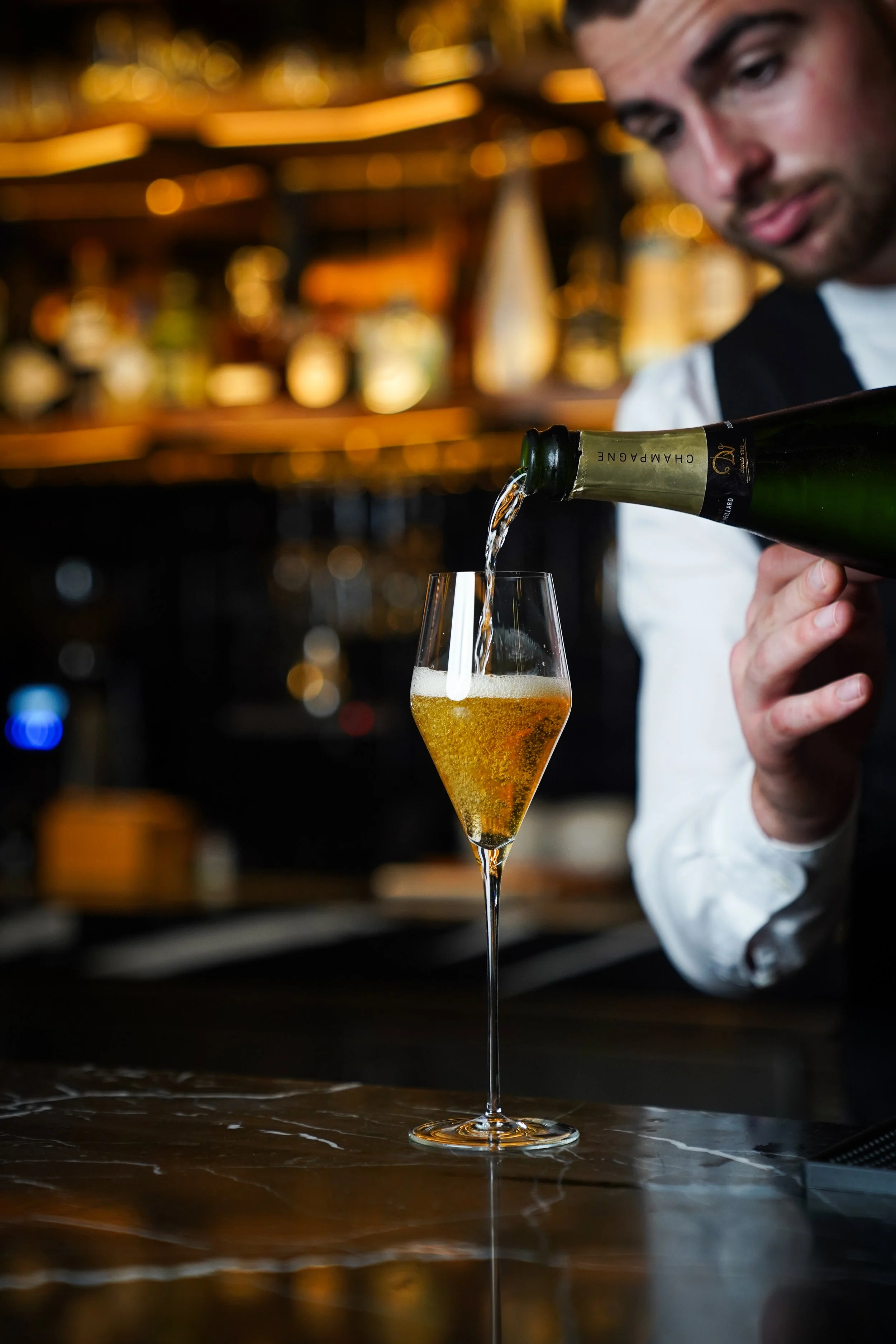 A bartender pours champagne into a champagne flute at a bar, with bottles and shelves in the background.