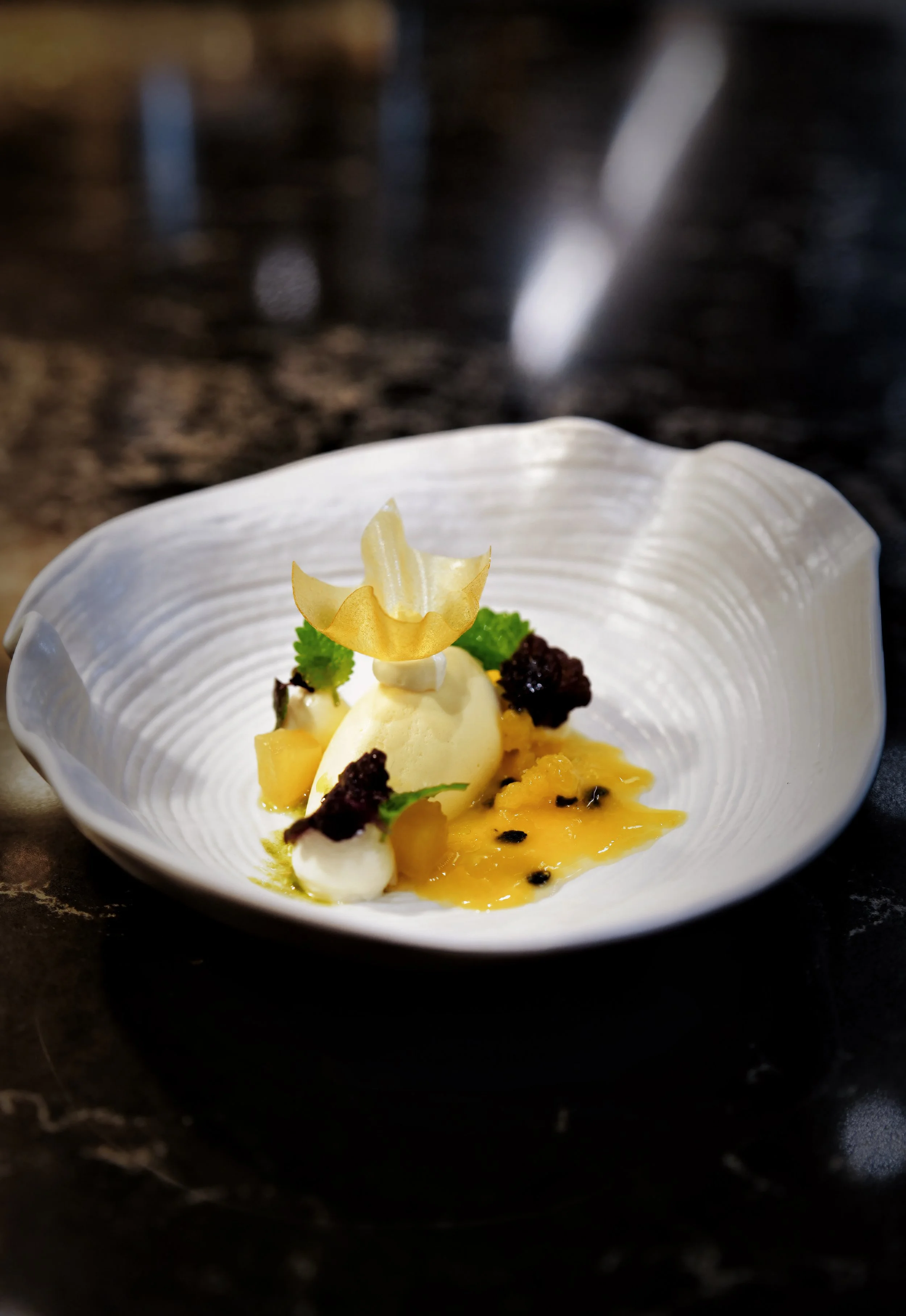 A plated dessert with ice cream, fruit sauce, candied fruit, mint leaves, and decorative edible flower on a white shell-shaped dish.