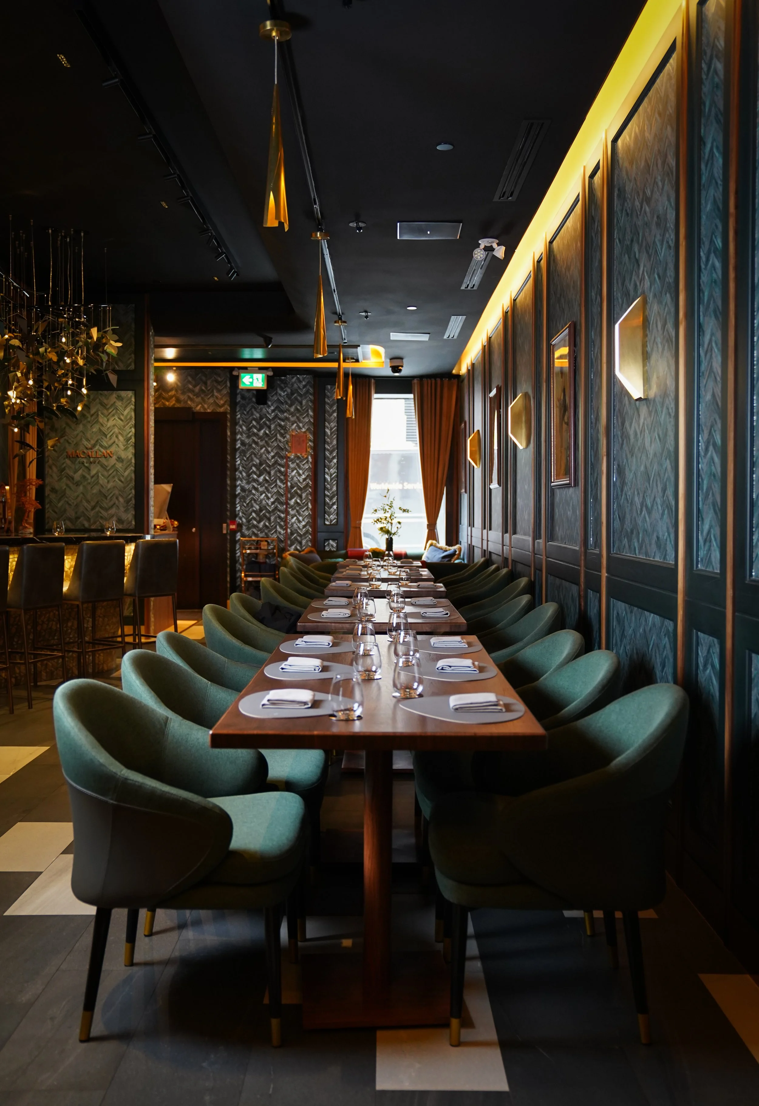 A long dining table set with place mats, cutlery, glasses, and napkins inside a modern restaurant, featuring dark wallpaper, green chairs, and warm lighting.