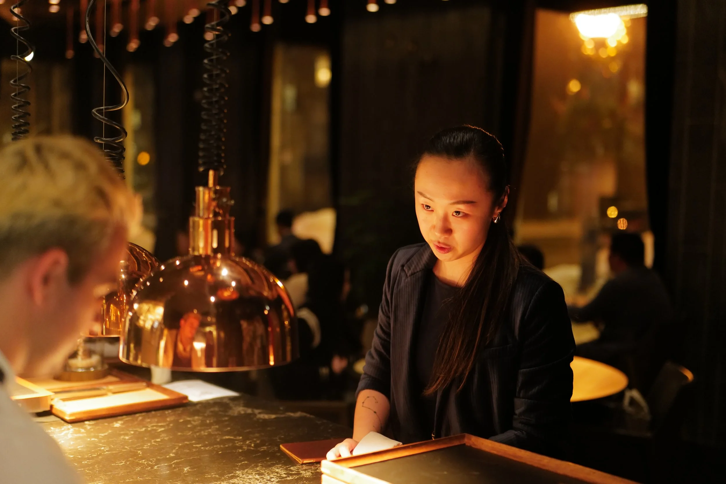 A woman working as a bartender at a dimly lit restaurant or bar, wearing a black jacket and listening to a patron, with copper pendant lights hanging above the bar.