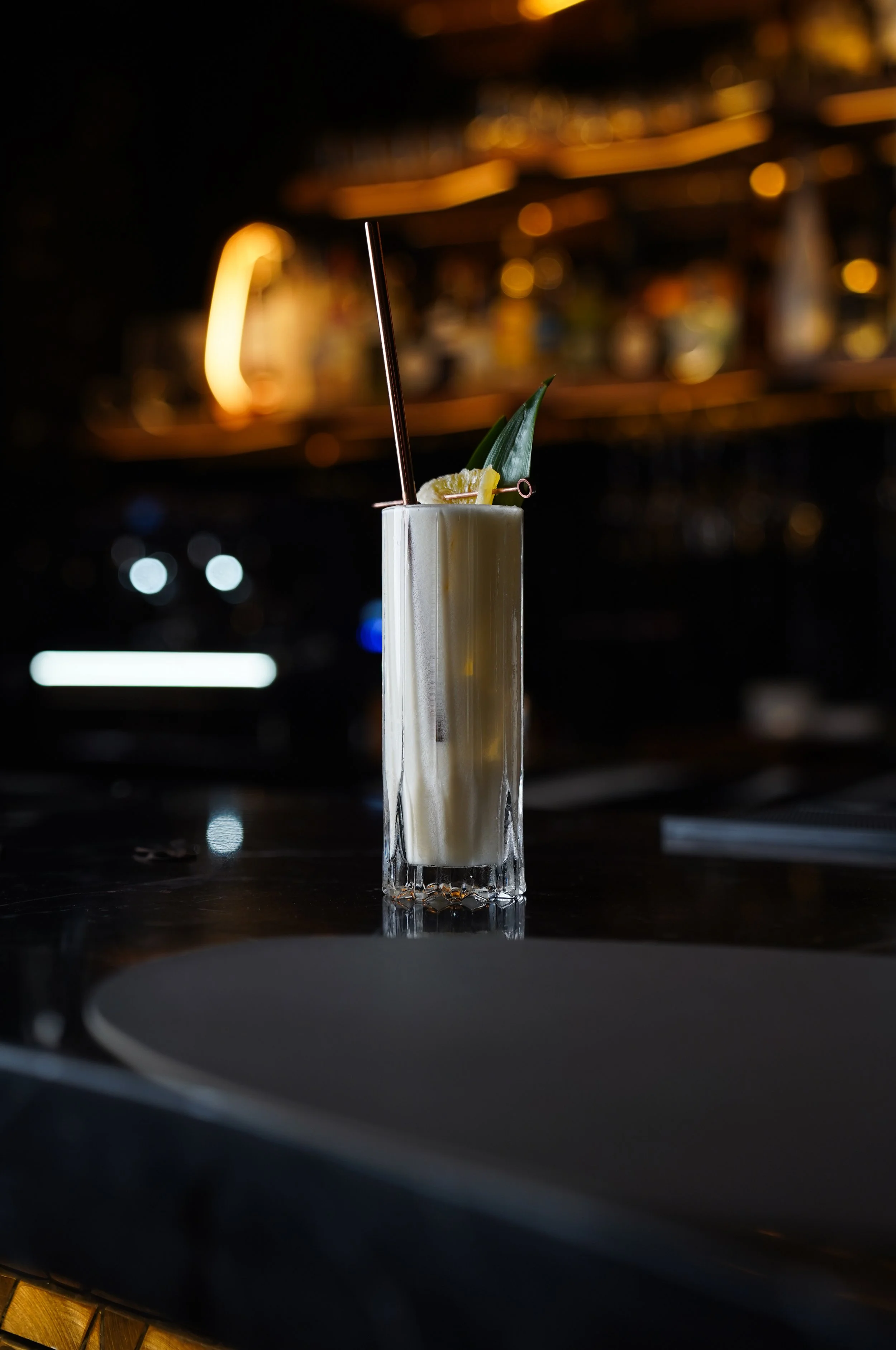 A creamy cocktail garnished with pineapple, lemon, and a leaf, served in a tall glass with a straw, on a dark bar counter.