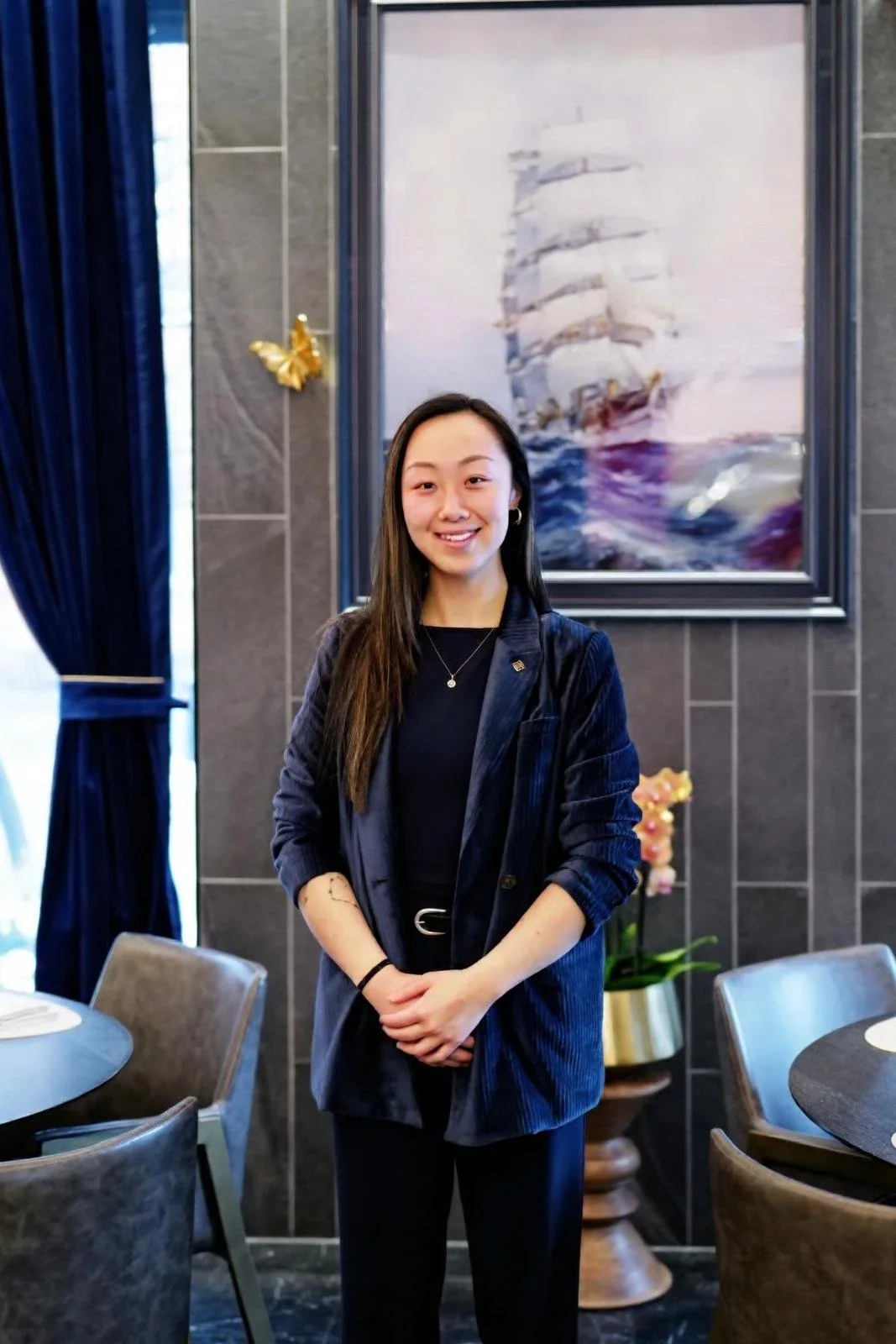 A young woman with long dark hair smiling, standing indoors in front of a painting of a sailing ship and a wall with dark tiles. She is wearing a dark blazer, black shirt, and black pants.
