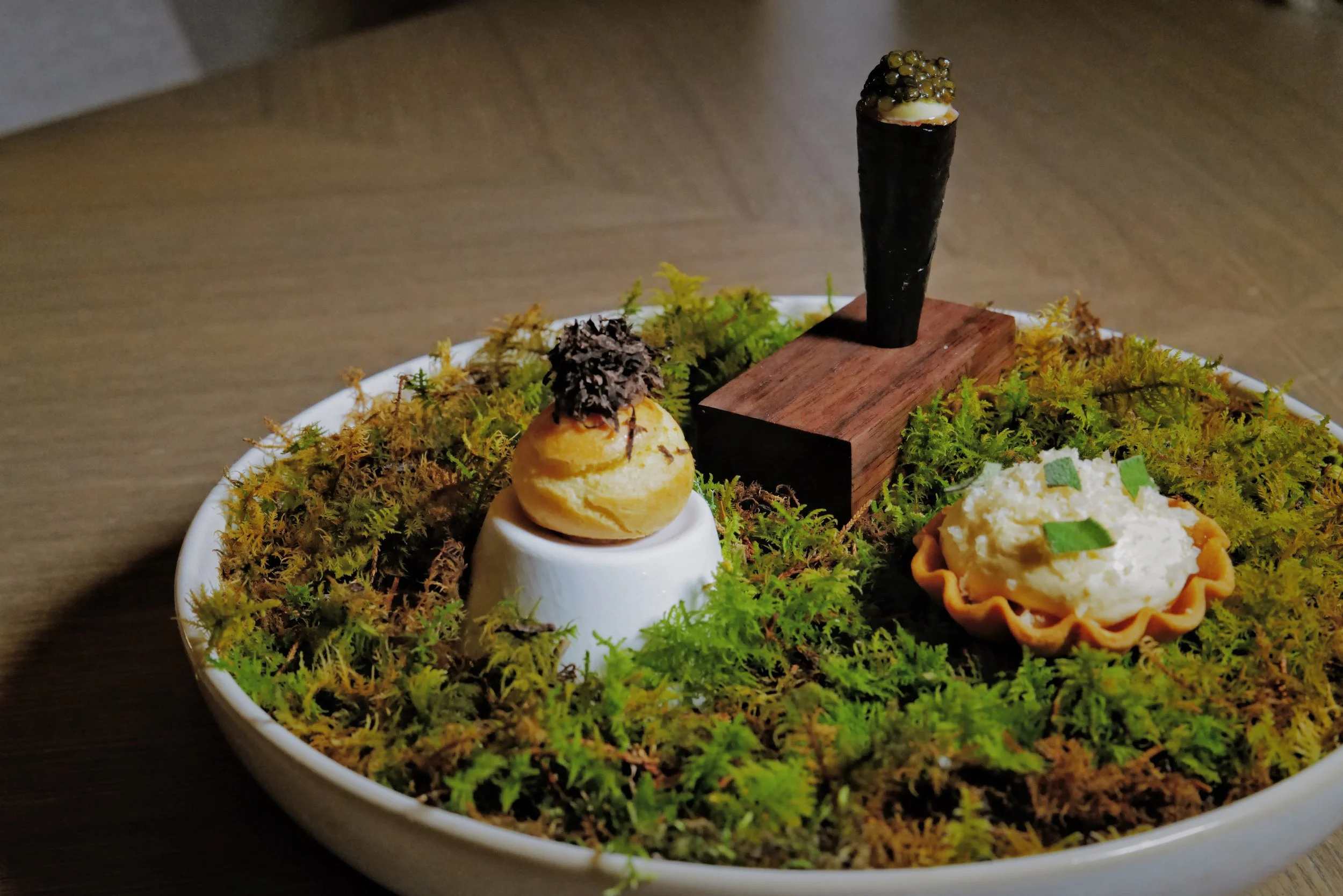 Miniature food display with moss, a small tart, a cream puff with chocolate shavings, and a chocolate cigar on a wooden stand.