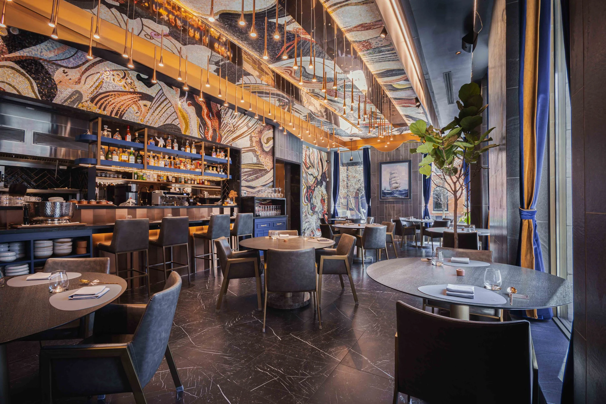 Elegant restaurant interior with round tables set with napkins and glasses, modern chairs, a bar with bottles on shelves, vibrant wall art, large windows with blue curtains, and a tall indoor plant.