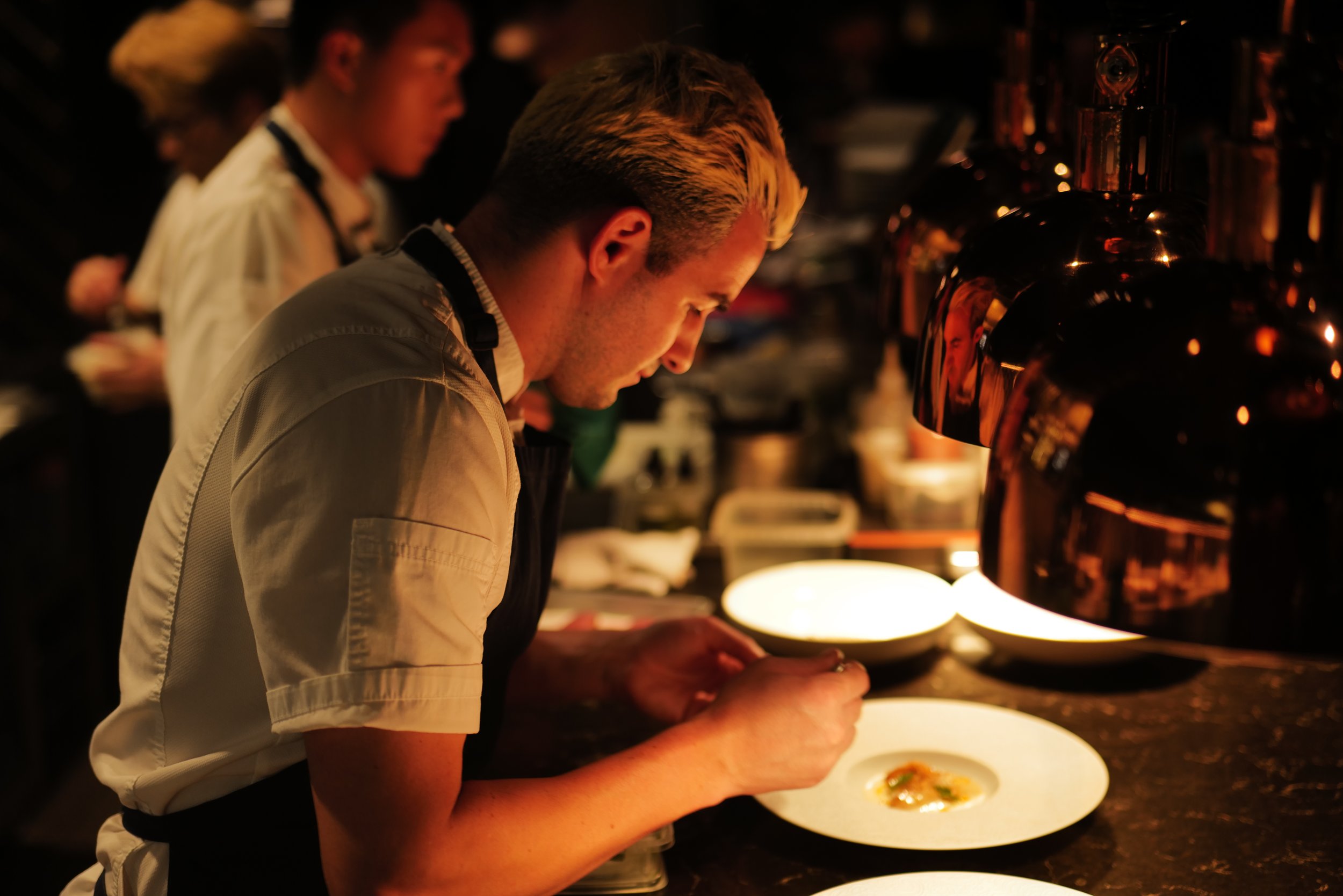 A chef plating a dish in a dimly lit kitchen with other chefs in the background.