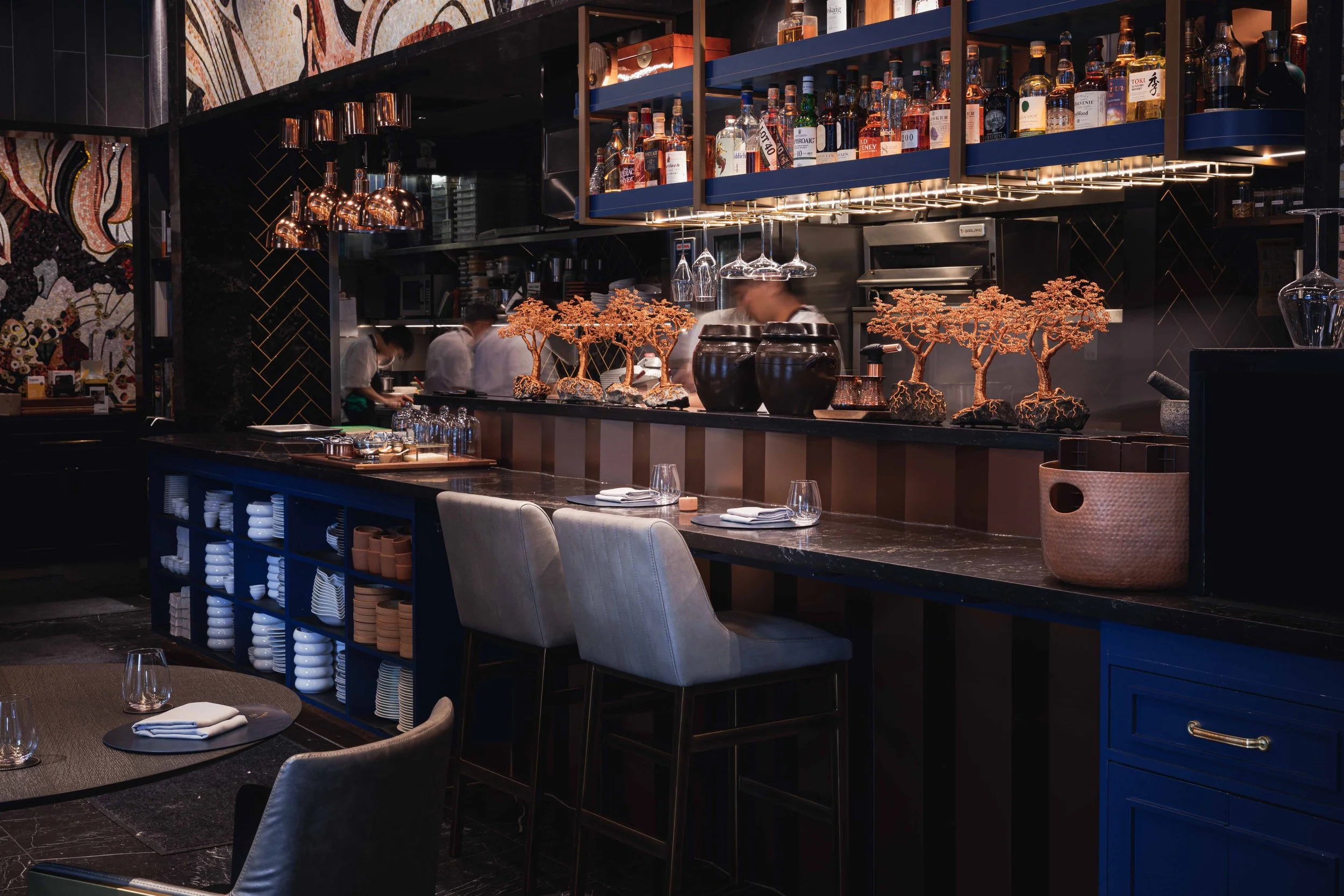 Interior of a modern restaurant bar with blue and black decor, shelves of liquor bottles, decorative trees, and a partially visible kitchen with chefs at work.