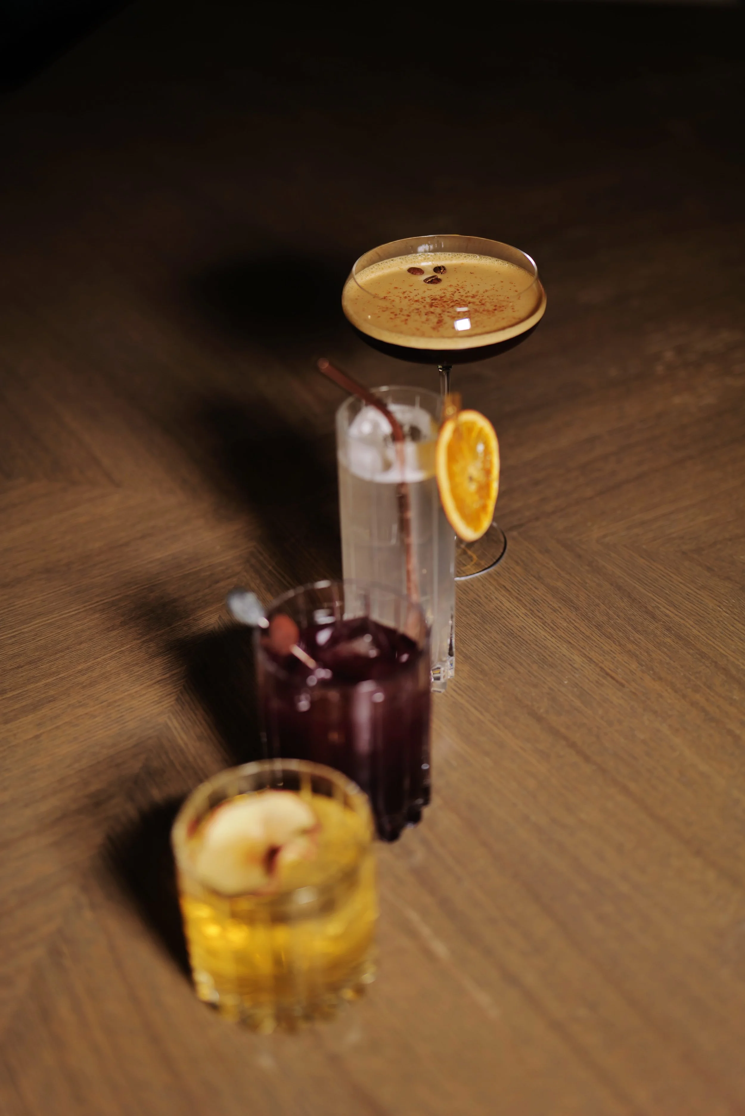 Three cocktails on a wooden surface: a coffee cocktail in a coupe glass garnished with cocoa powder, a tall glass with dark liquid and a slice of orange, and a whiskey drink with ice and lemon slice.
