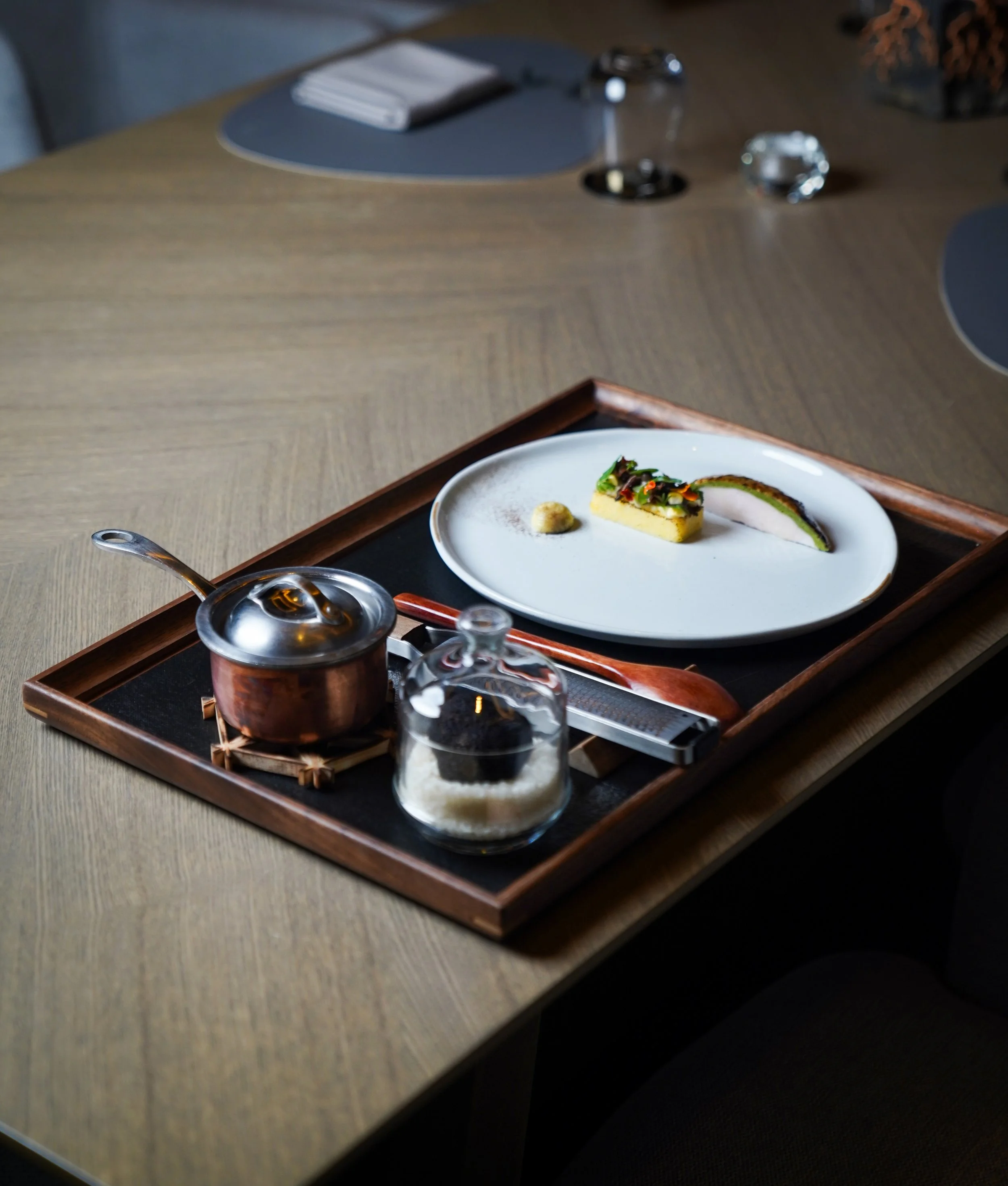 A wooden tray on a dining table holds a white plate with a small serving of food, a glass jar with sugar, a copper pot with a lid, a pair of chopsticks, and a fork. The table has a dark surface with a placemat in the background.