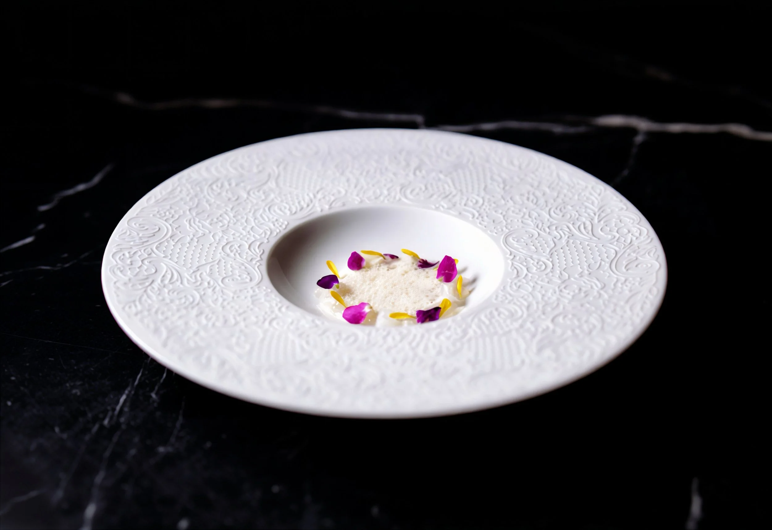 A white, textured plate with intricate designs around the rim, containing a small amount of white powder garnished with purple and yellow flower petals, placed on a black marble surface.