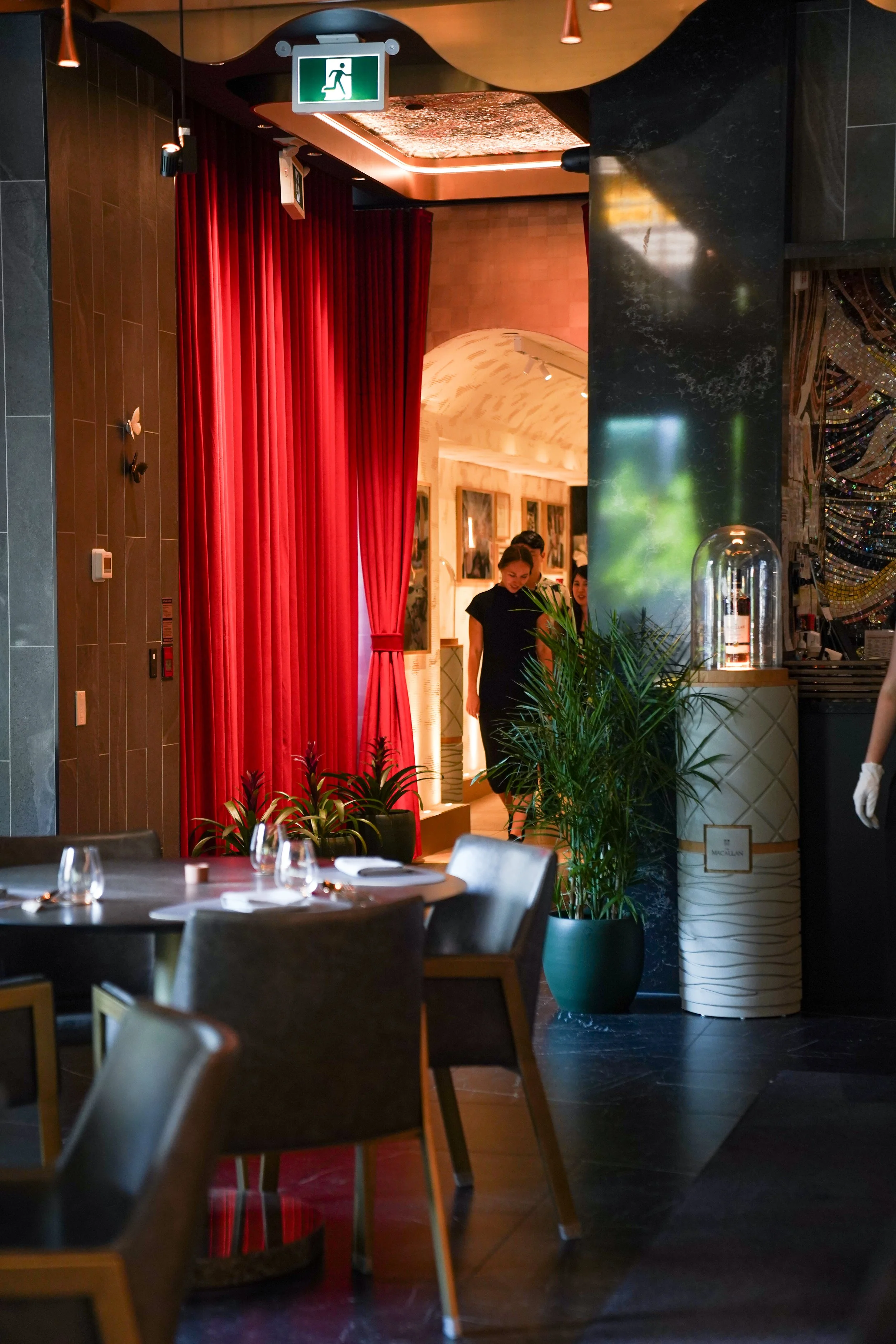 Dining area in a restaurant with tables set for guests, a large green plant near a black marble pillar, and red curtains. People are walking in the background near framed photographs, with an emergency exit sign hanging from the ceiling.
