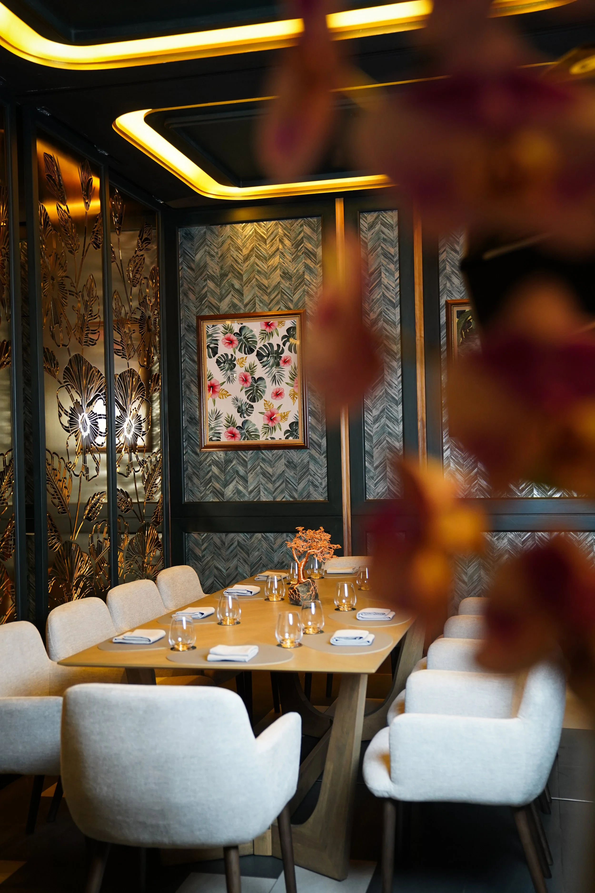 A fancy dining room with a wooden table set with white napkins and glasses, surrounded by beige upholstered chairs. The walls feature dark patterned wallpaper, framed floral artwork, and gold decorative panels with a butterfly motif. A branch with pi