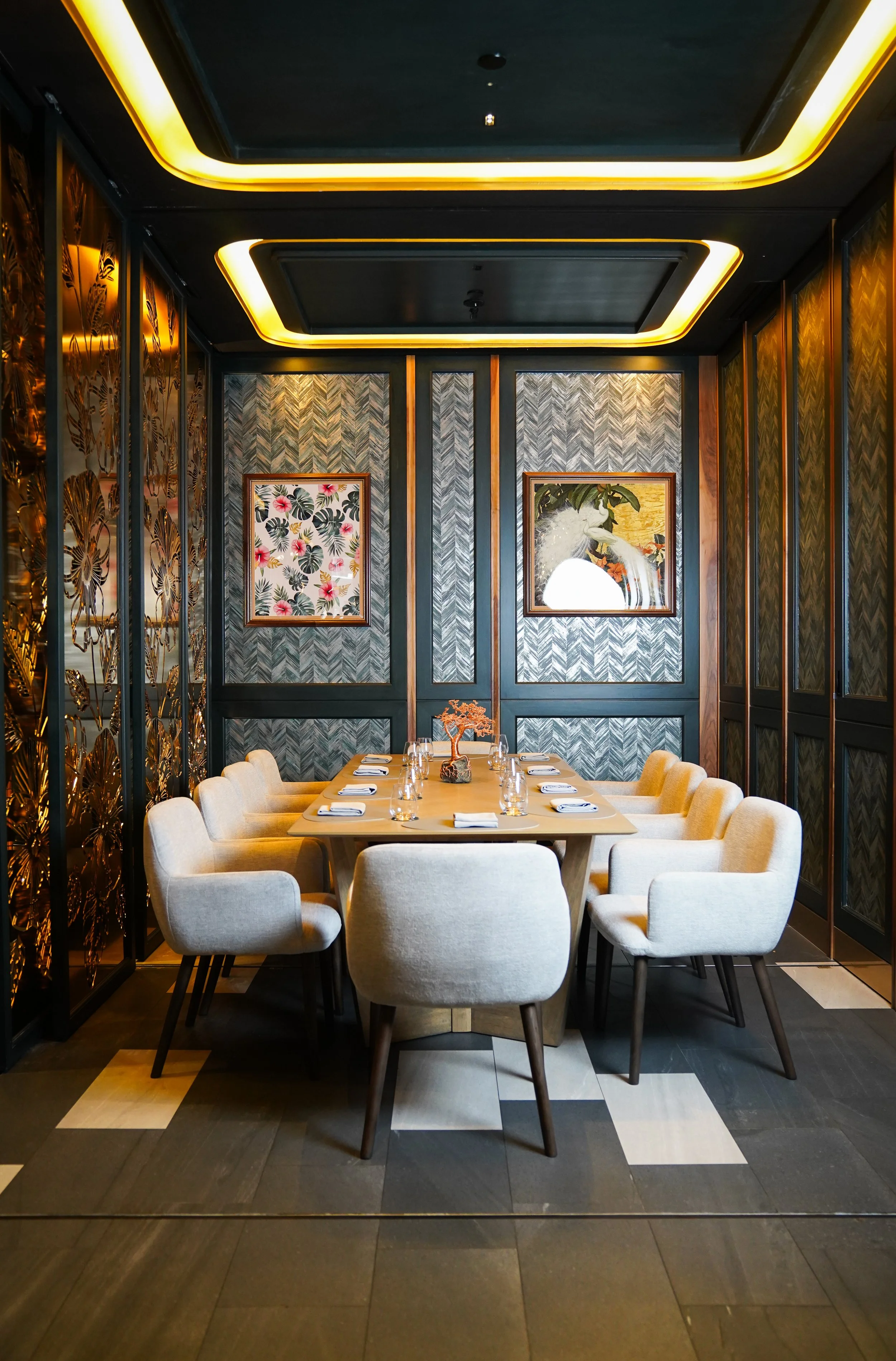 A formal dining room with a rectangular table set for eight, surrounded by beige upholstered chairs. The walls feature dark and patterned artwork, and there are decorative panels with floral and leafy designs on the side walls. The ceiling has recess