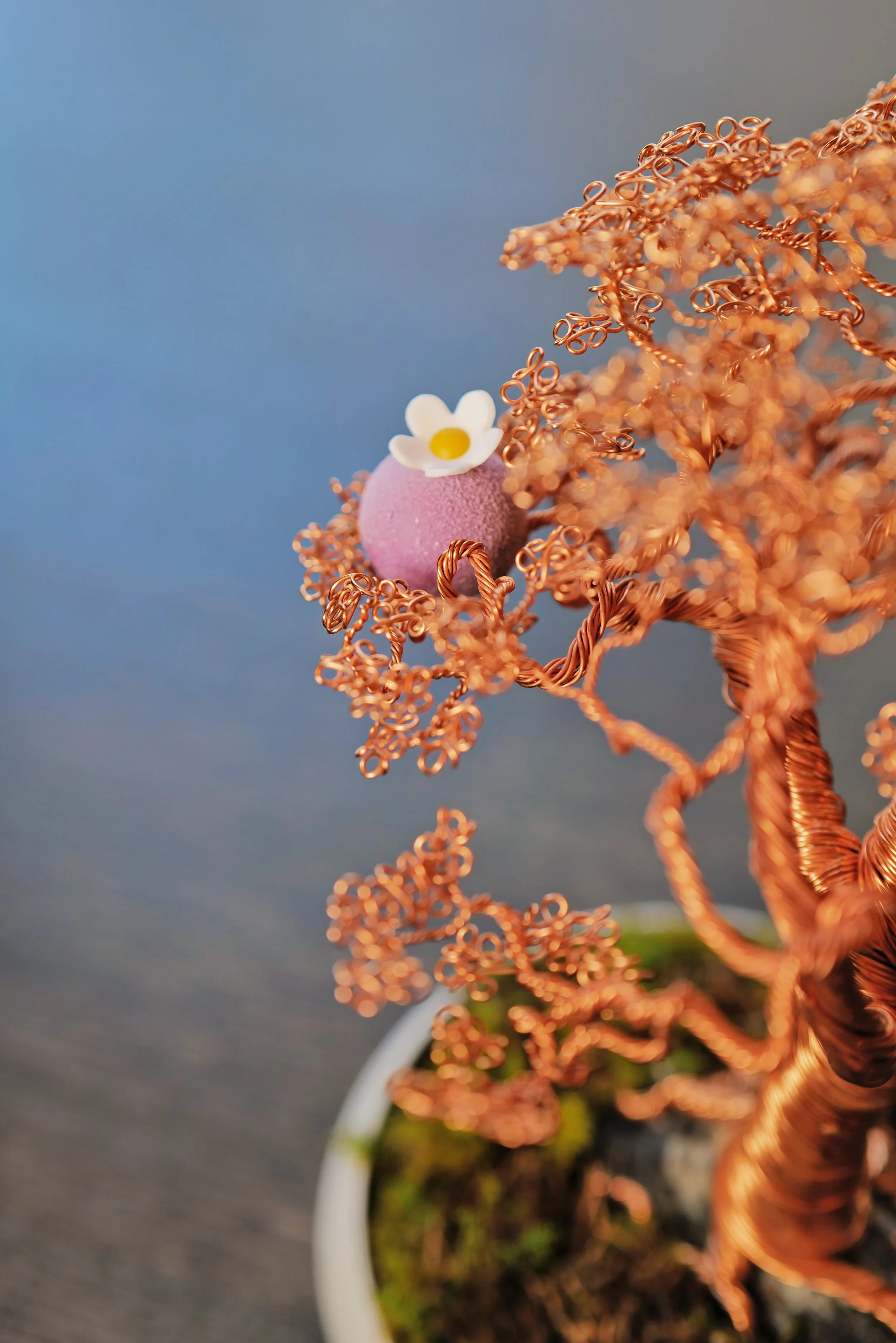 A miniature copper wire tree with intricate twisting branches in a white pot, decorated with a small pink foam ball and a tiny white daisy flower on top.