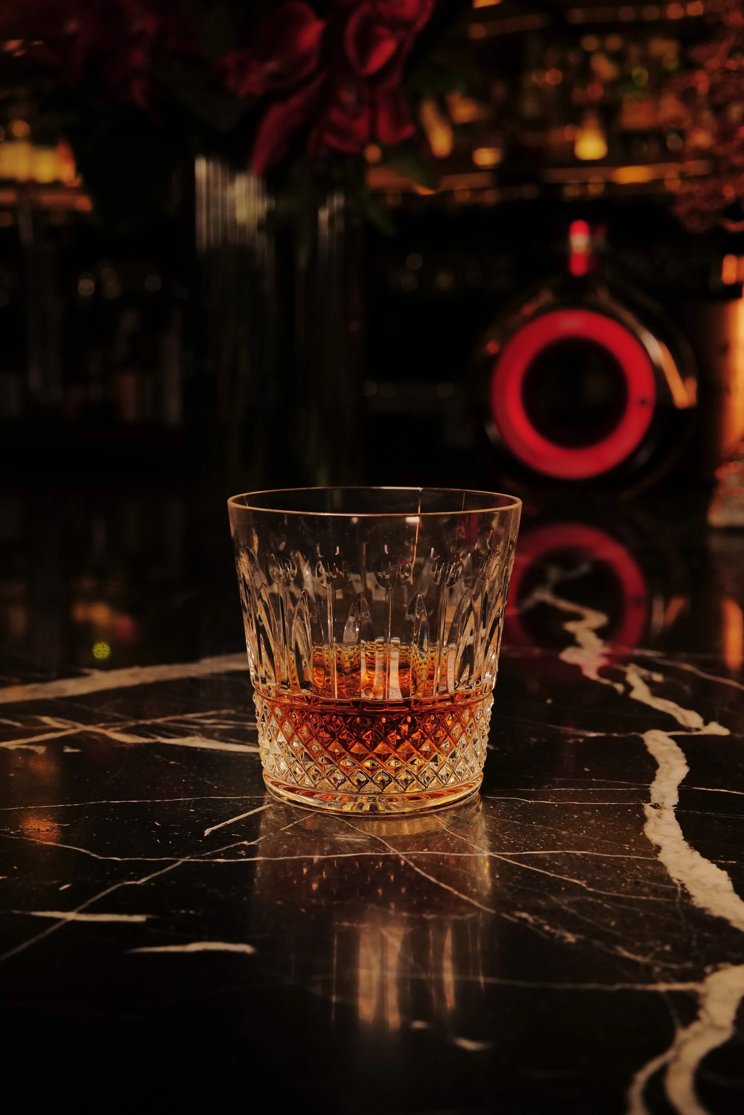 A glass of amber liquor on a black marble table with white veins, reflections visible on the surface.