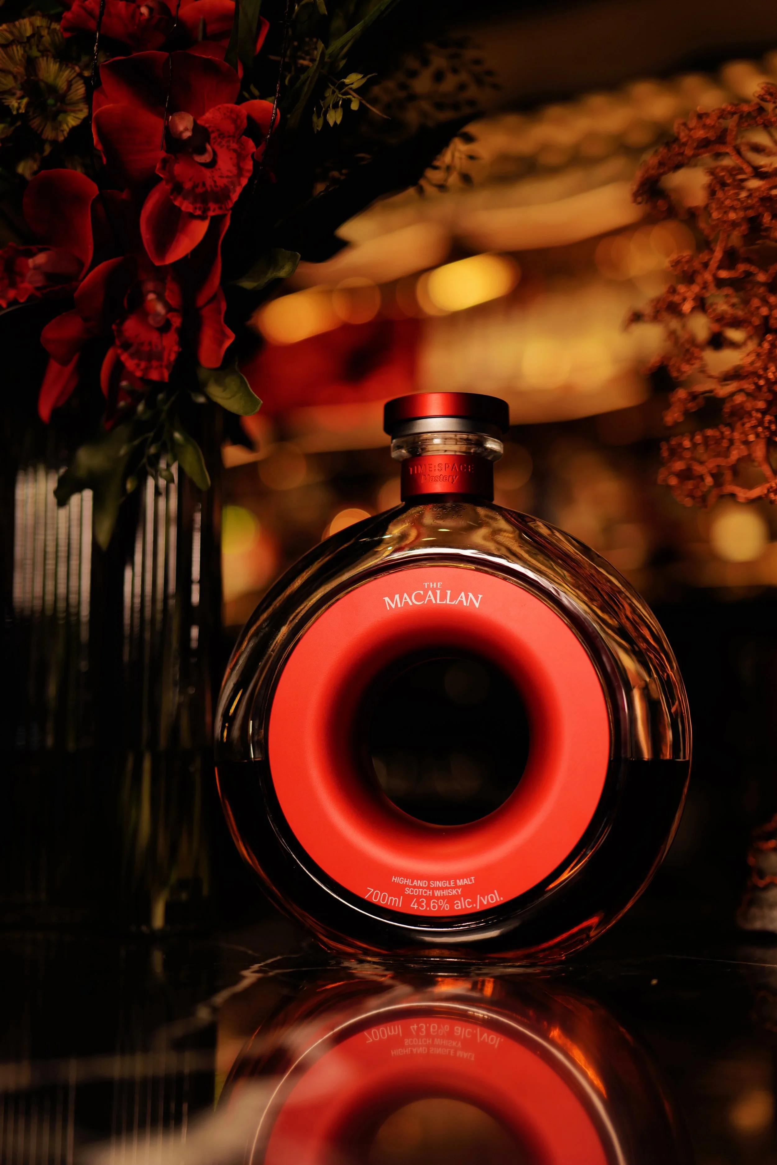A bottle of The Macallan Highland Single Malt Scotch Whisky with a red circular design on the label, placed on a reflective surface surrounded by dark floral arrangements with red flowers, giving a warm, ambient lighting effect.