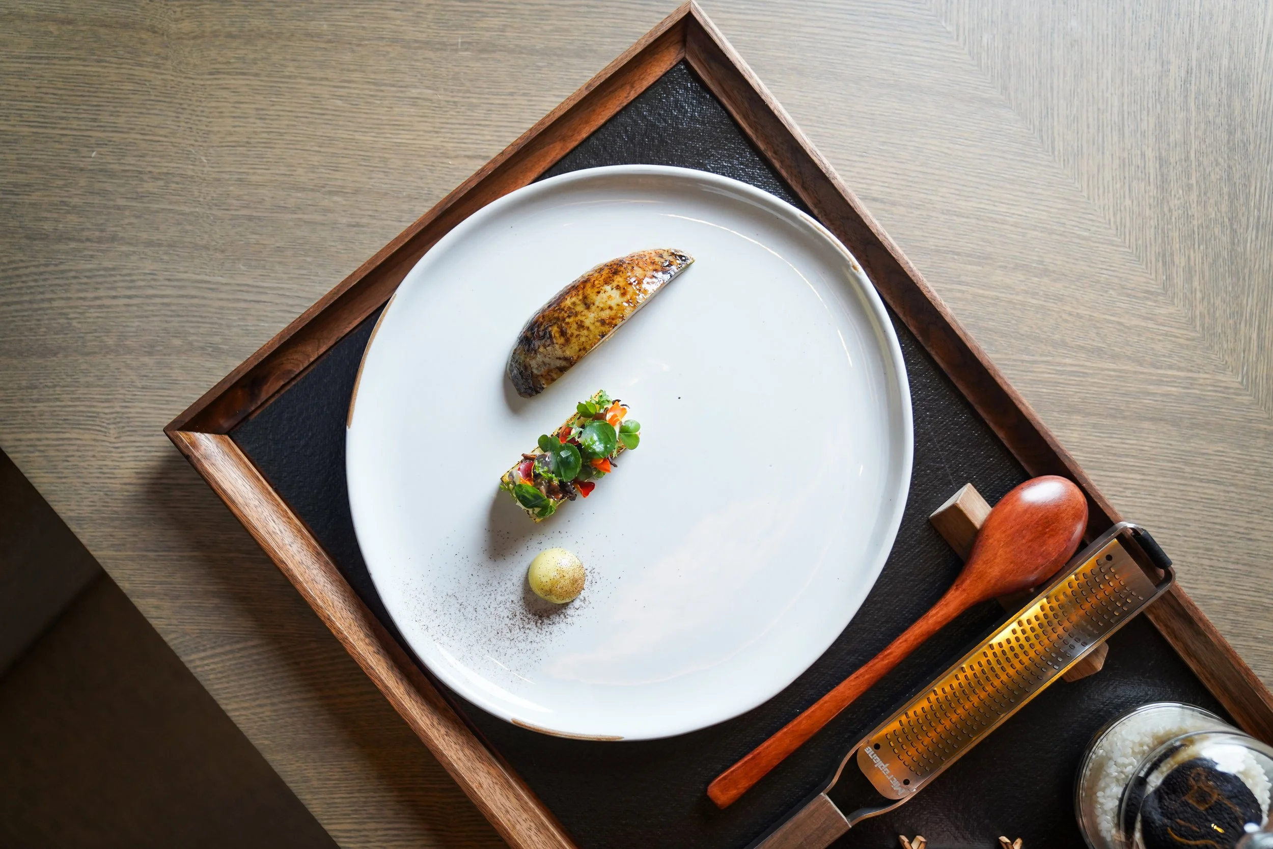 A minimalist plate with a roasted banana, a small colorful vegetable terrine, a dollop of white sauce, and a light dusting of powder, served on a round white dish on a wooden tray with a wooden spoon and a small jar of shaved ice.