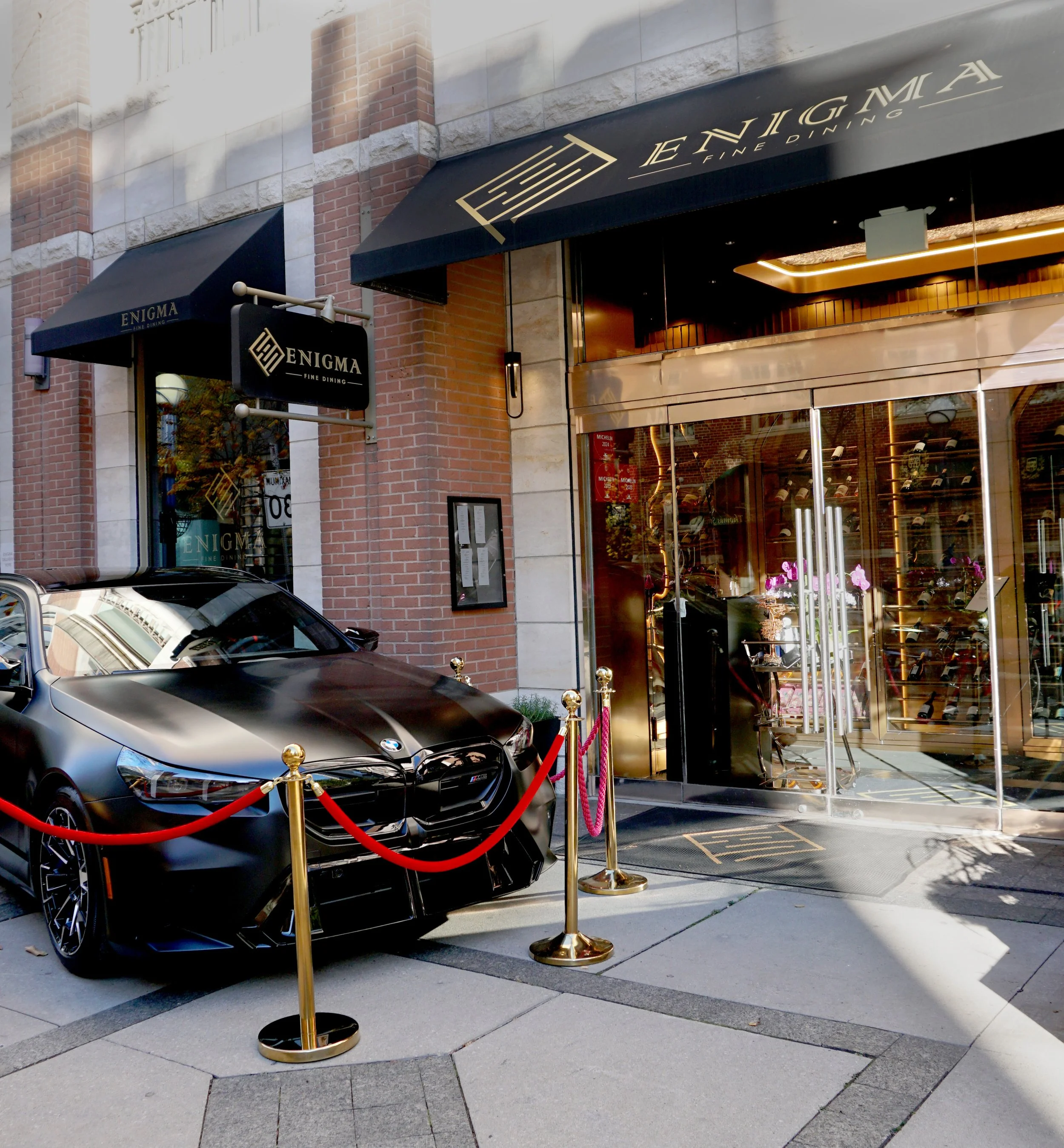 A black BMW sports car parked outside ENIGMA Fine Dining restaurant, which is marked by a black canopy and a sign; the car is roped off with red velvet ropes and gold stanchions at the entrance.