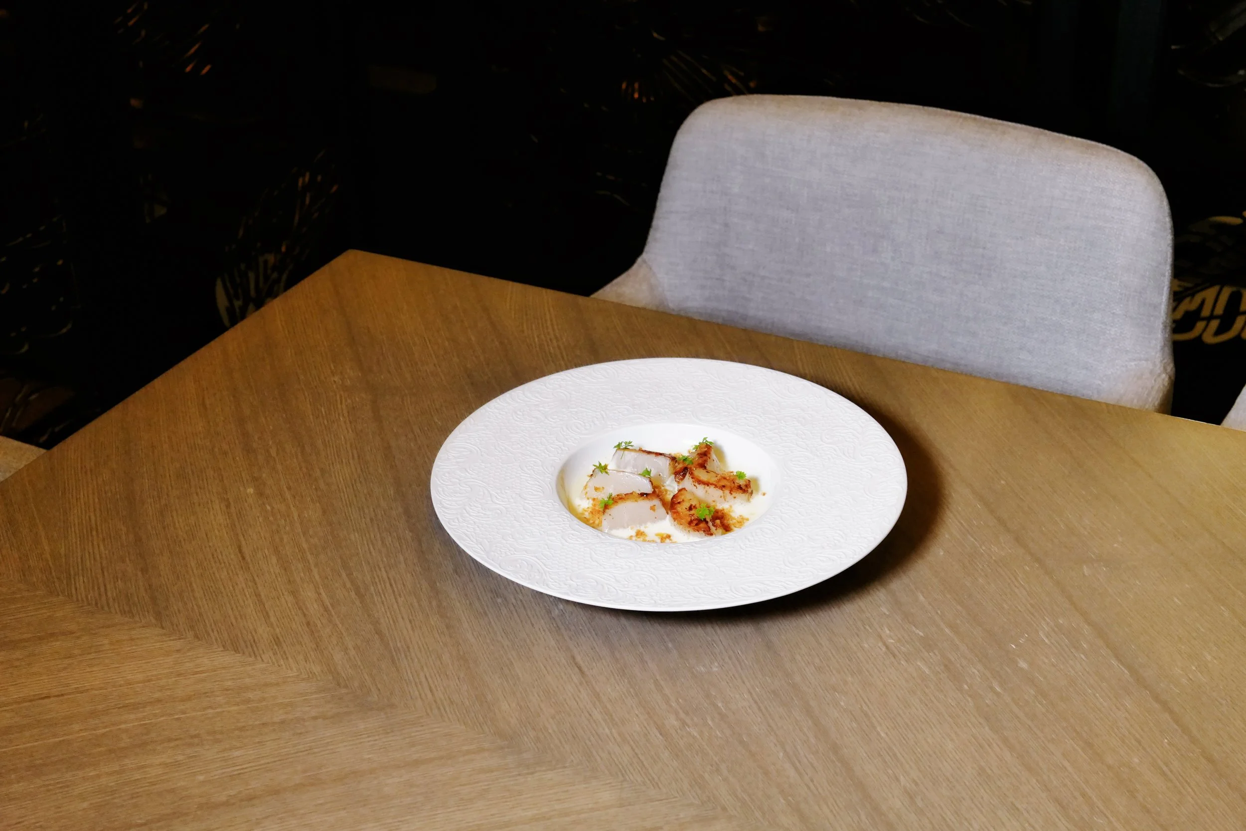 A white decorative plate with a dish of sliced cooked meat garnished with herbs on a wooden table, with a light-colored upholstered chair nearby.